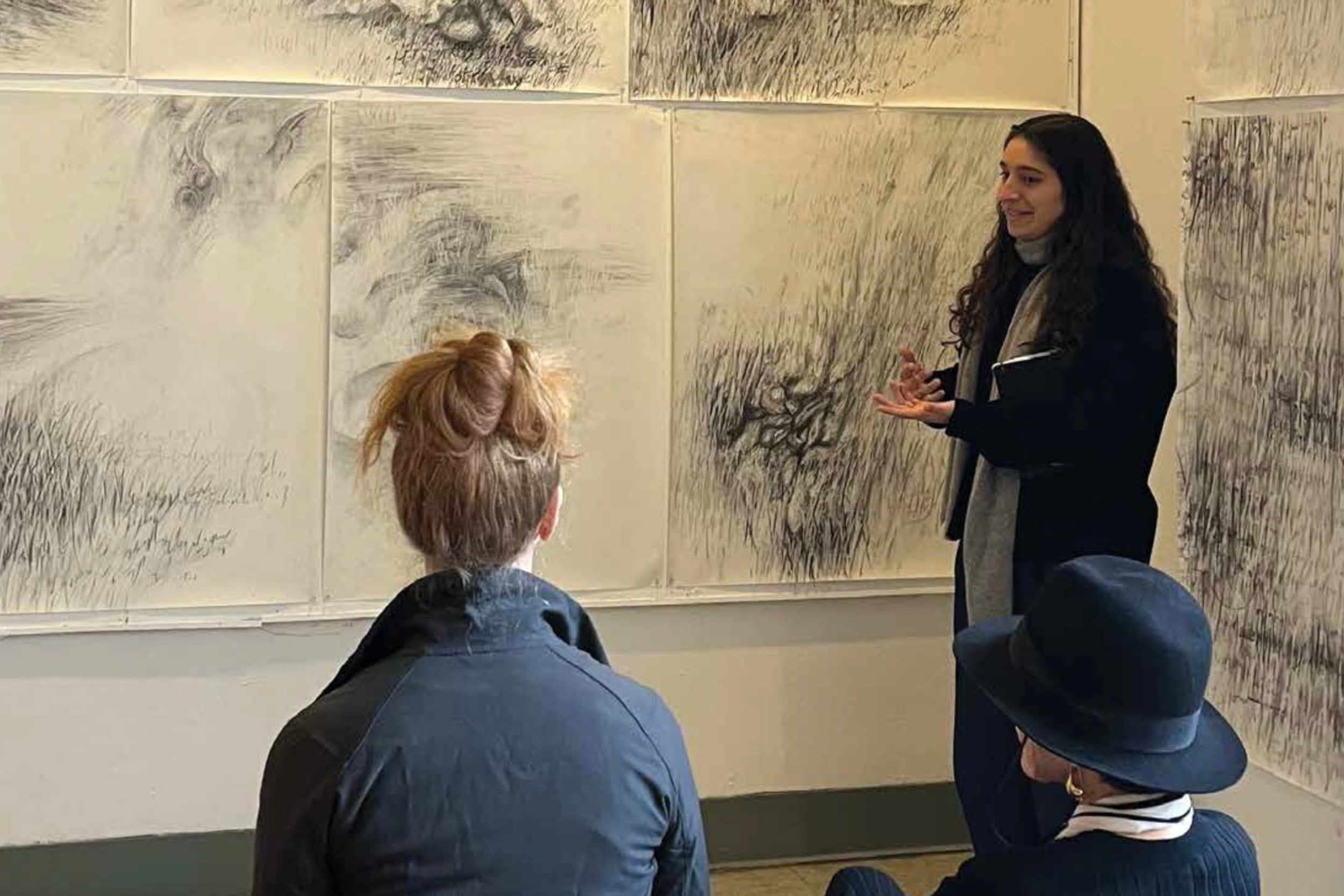 A young woman stands in front of a wall adorned with large black and white sketches. She's speaking to two people seated in chairs in front of her. 