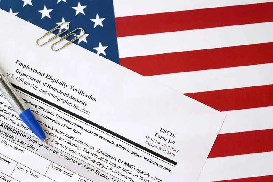 US employment eligibility verification form on a table, with American flag in the background, a silver and blue pen, and paper clips.