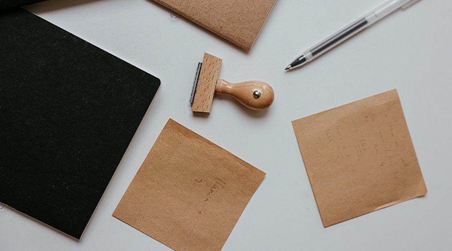 Stationery items including black and brown notebooks, a wooden-handled stamp, a silver pen, and two sticky notes with handwritten notes on a white surface.