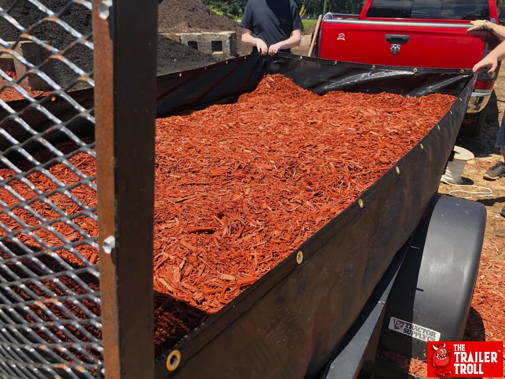 Close-up of a full utility trailer load of red mulch secured by a black trailer liner with strong grommets for safe transport of landscaping supplies.