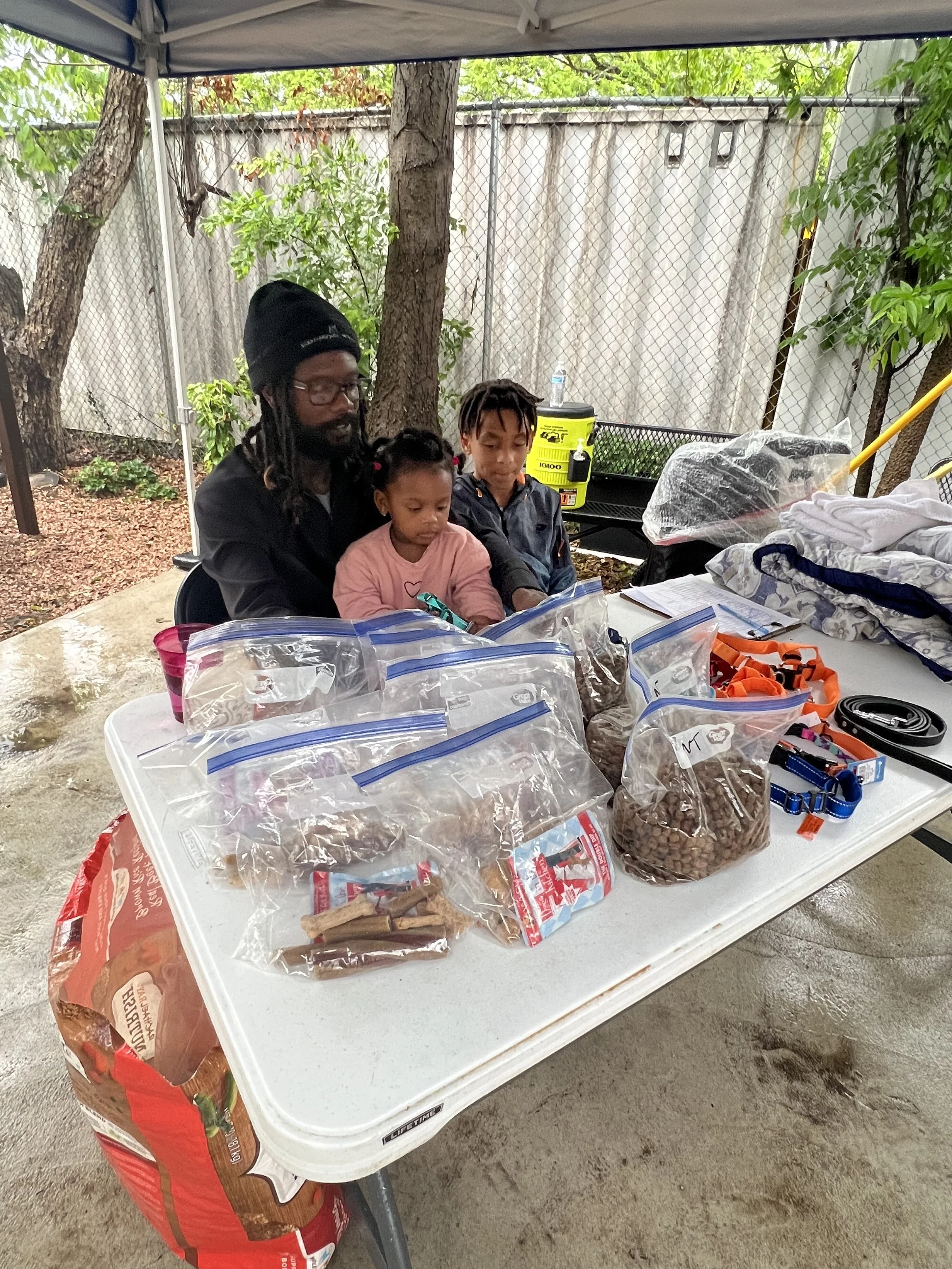 Three children and a man sitting at a table outdoors under a canopy, with bags of pet food, dog collars, and pet accessories on the table.
