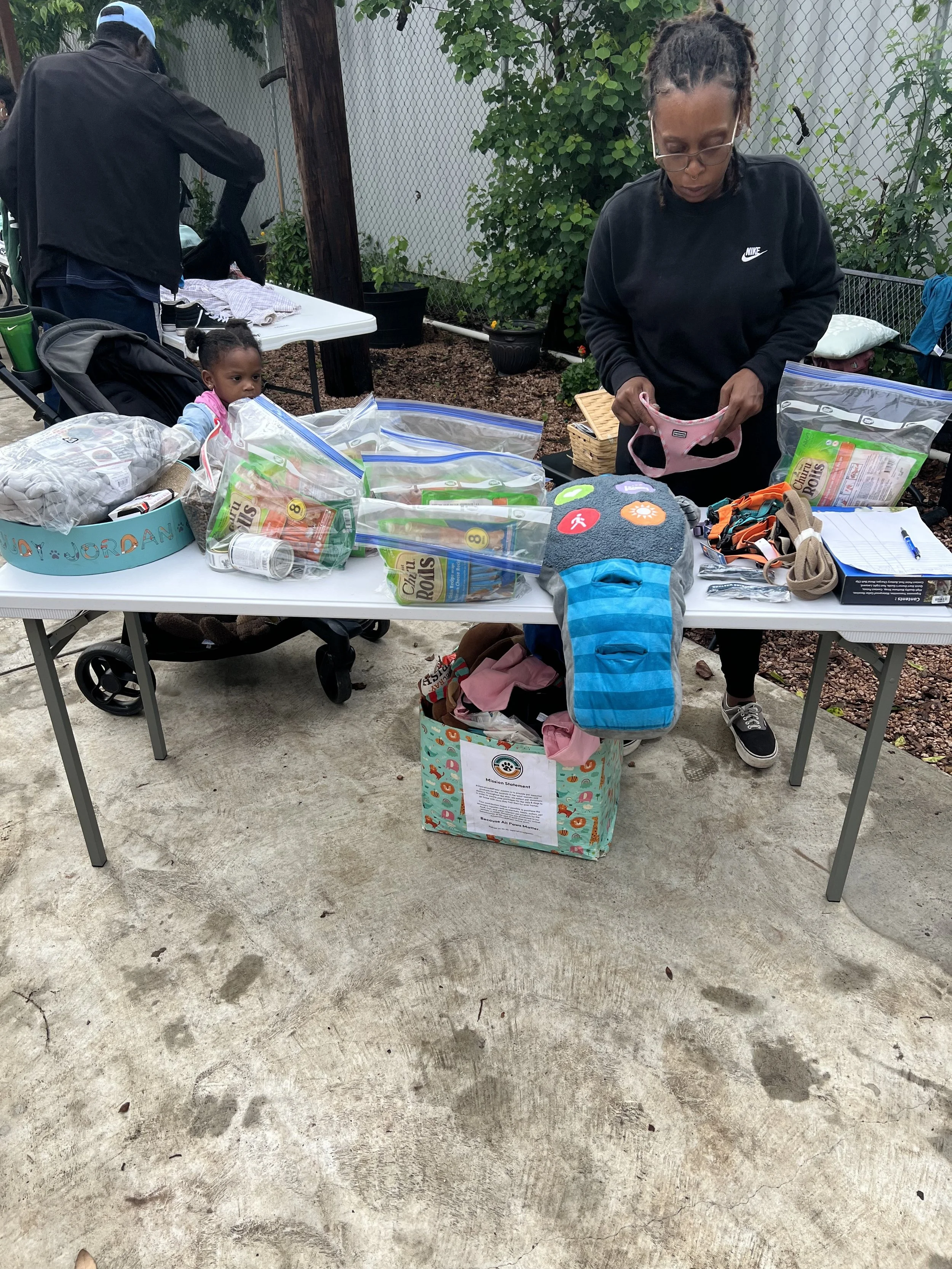 People setting up a table with various bags of supplies and a box, with a woman organizing items and a child sitting behind the table.