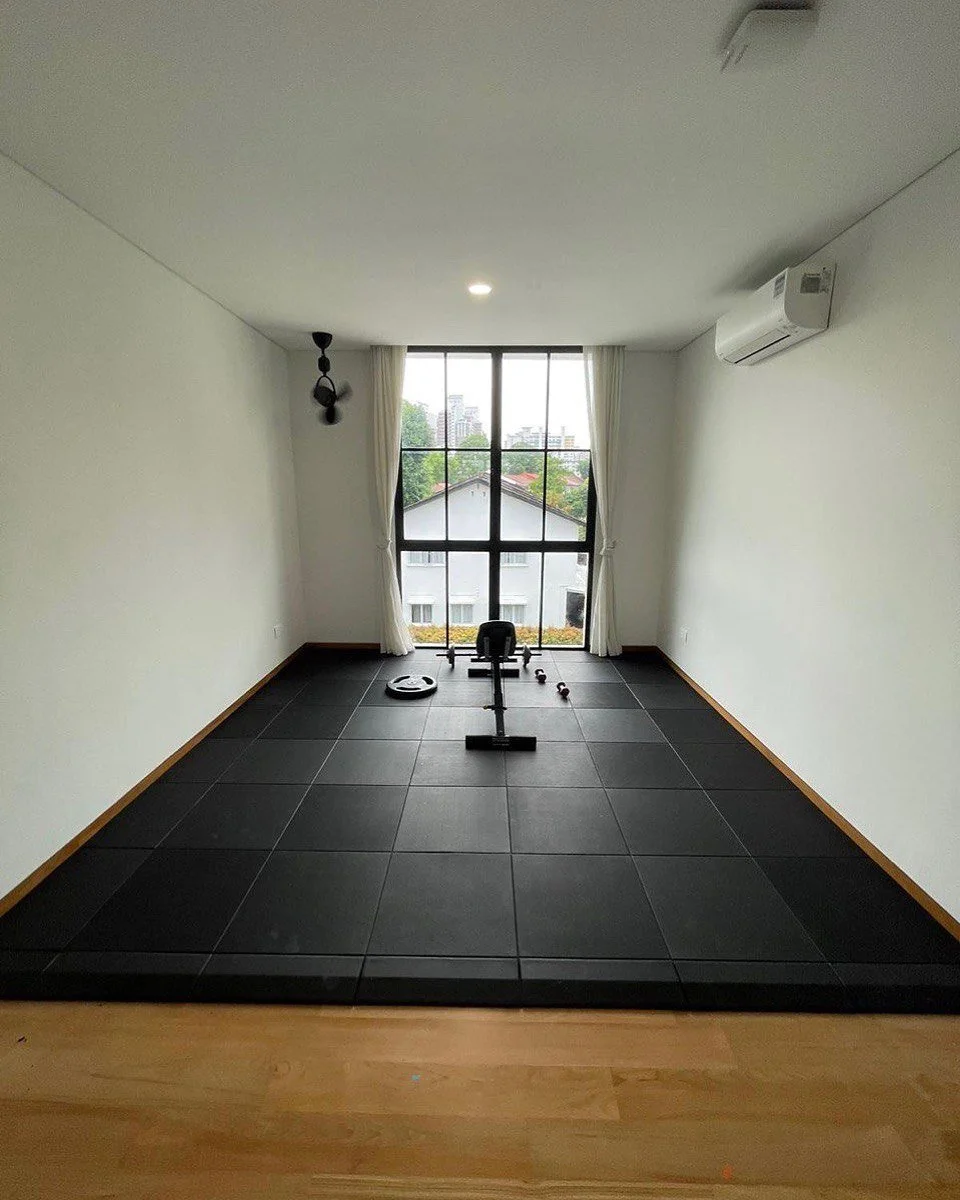 Empty room with black exercise mats, a rowing machine, a weight, and a roller, facing a large window with white curtains.