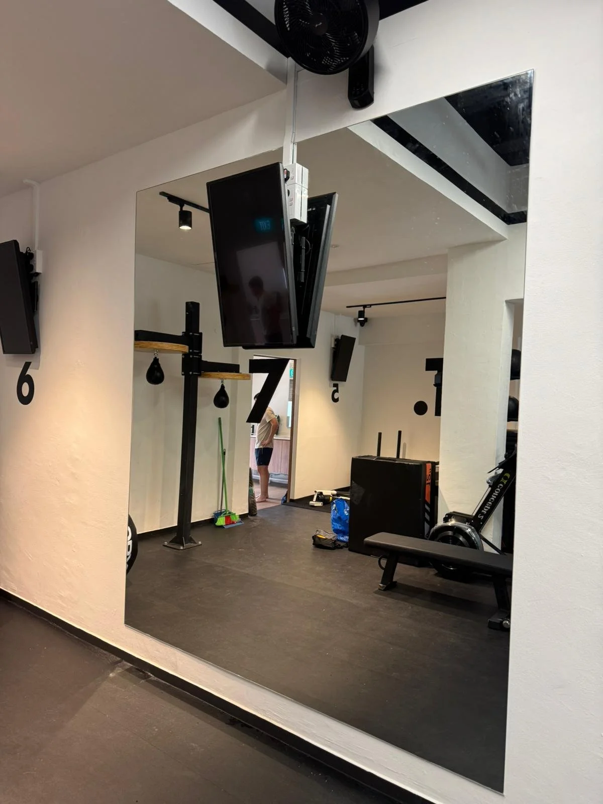 A gym with workout equipment, a large mirror, and a digital timer showing 19:02. There are wall-mounted TVs and a person partially visible in the doorway.