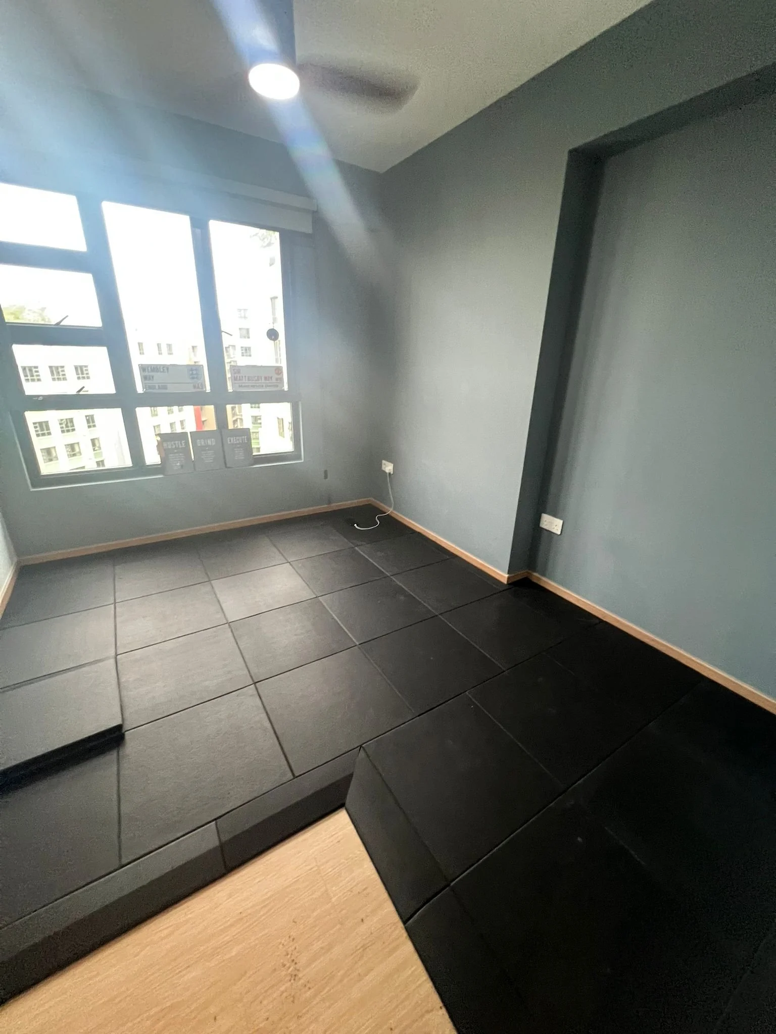 Empty room with large window, black tile flooring, gray walls, ceiling fan with light, and electrical outlets.