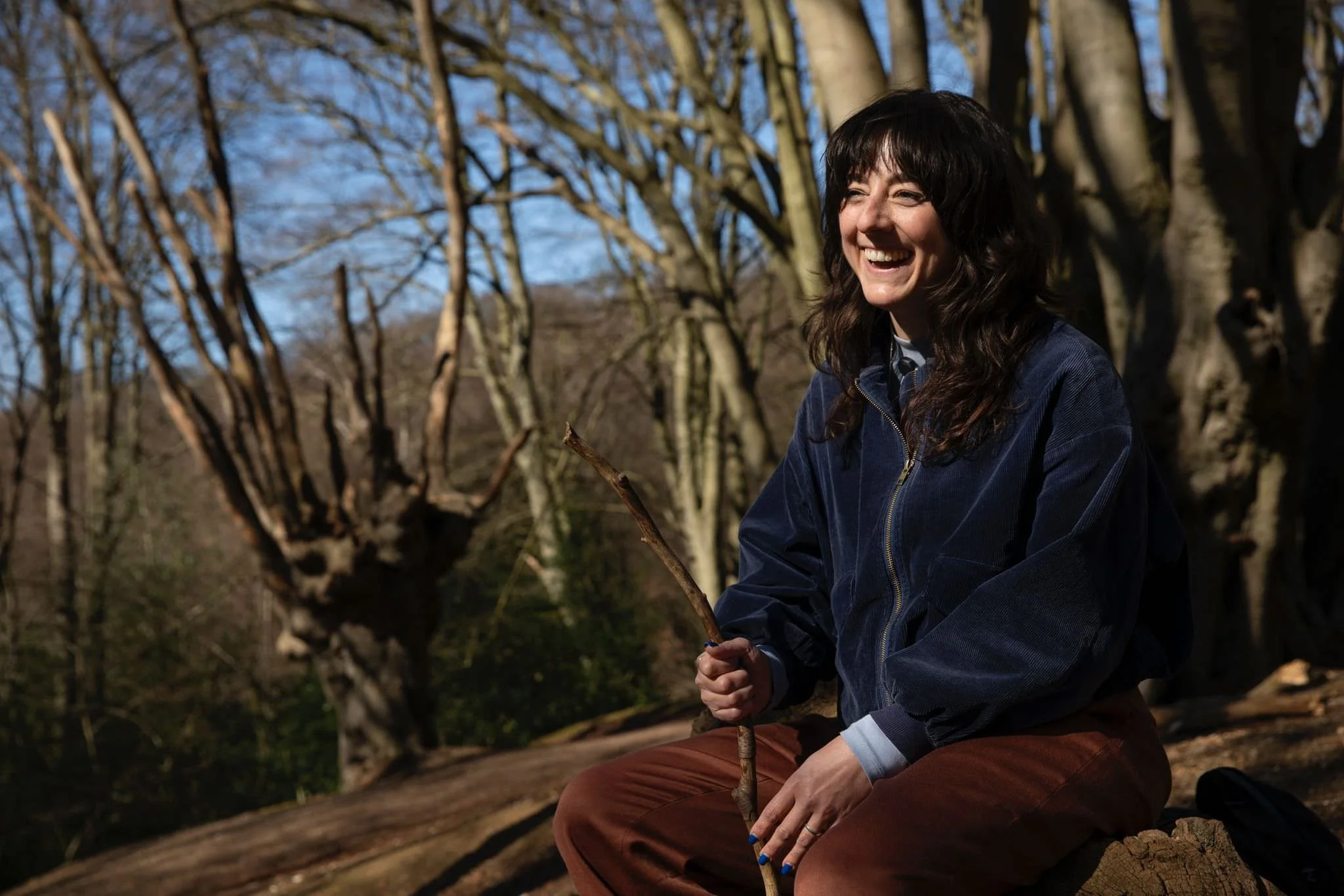 Jess Jorgensen sits, smiling during a Field Trip in England's Epping Forest, on a fallen tree stump in the forest. It's a sunny day. She's holding a good stick.