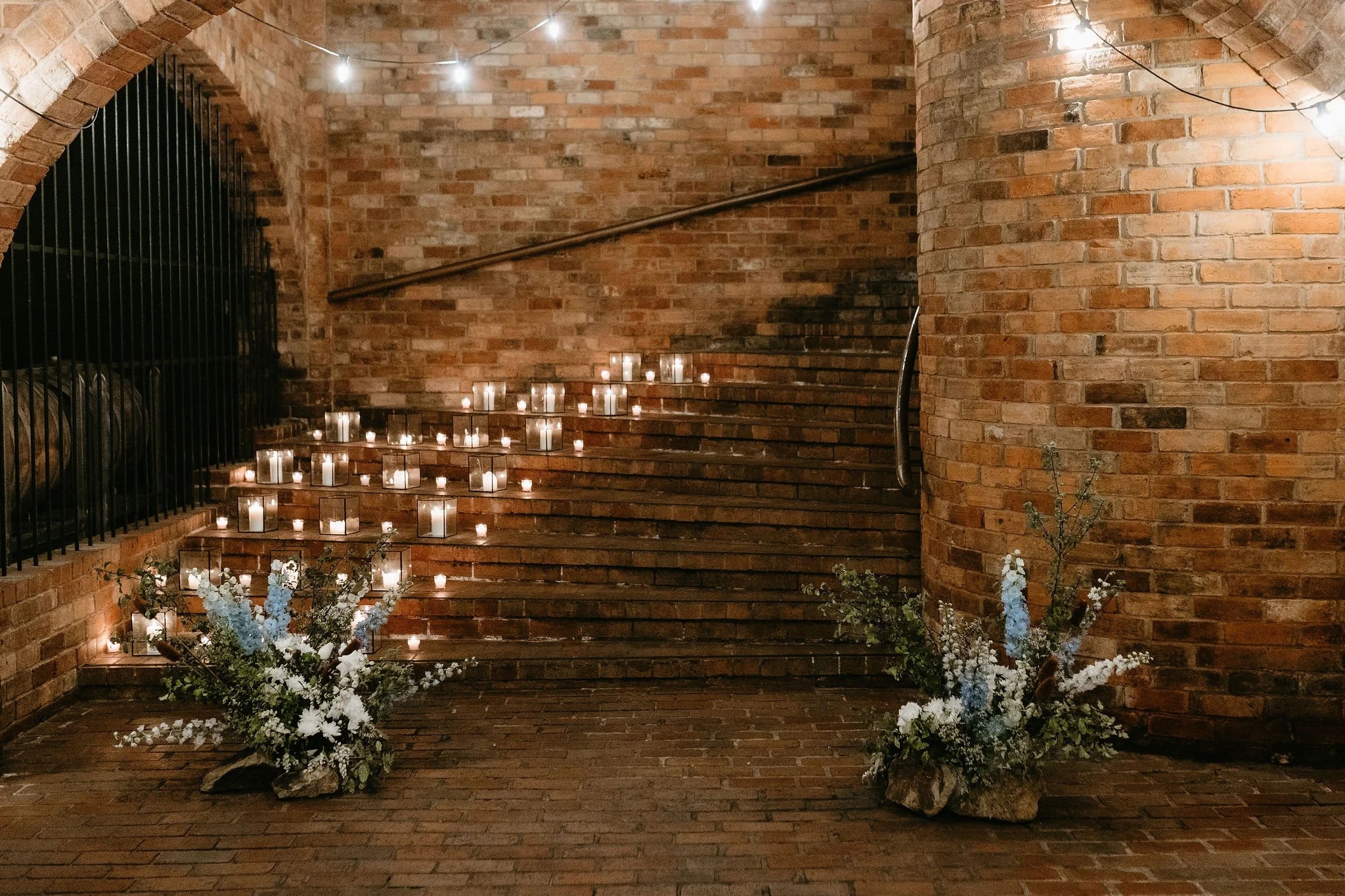 Brick staircase decorated with numerous lit candles in glass holders, and floral arrangements with white and light blue flowers at the base.