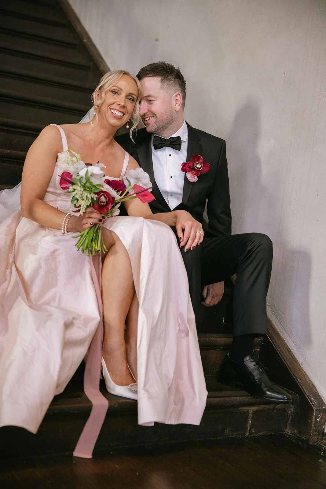 Bride and groom sitting on stairs, smiling, with the bride holding a bouquet, bride in pink dress and white heels, groom in black tuxedo with bow tie