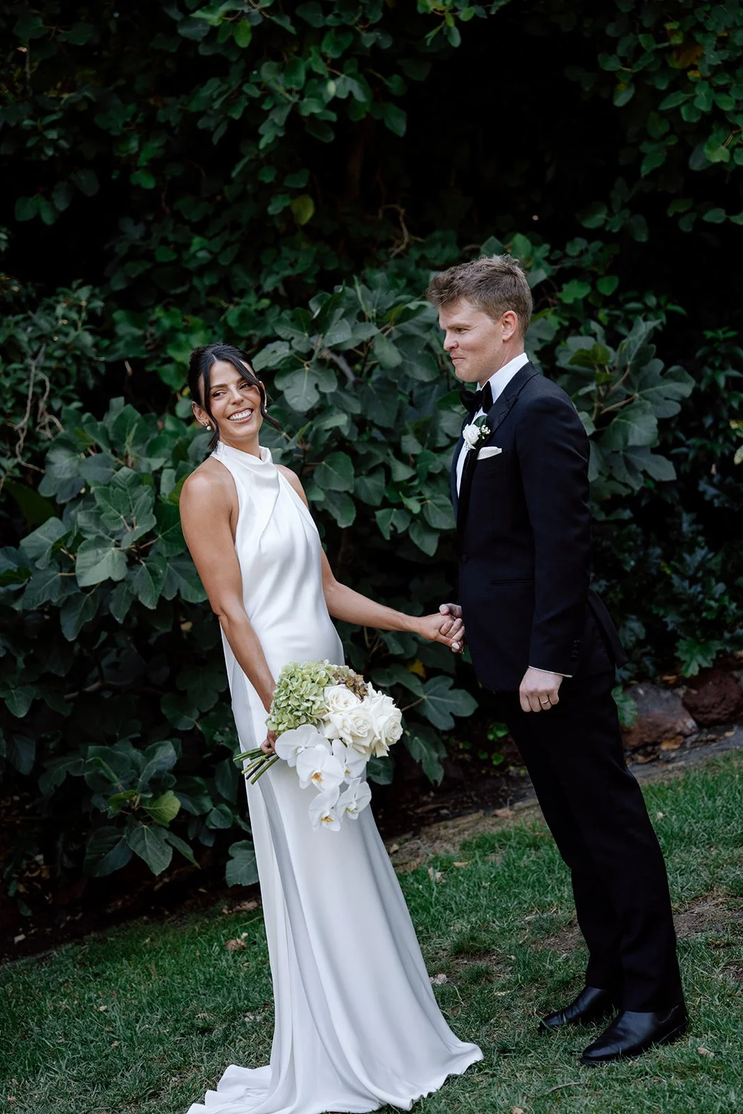 A bride and groom standing outdoors on grass, holding hands, with large green leafy plants behind them. The bride has dark hair in an updo, wearing a sleeveless white wedding dress and holding a bouquet of white flowers. The groom has short hair, wea