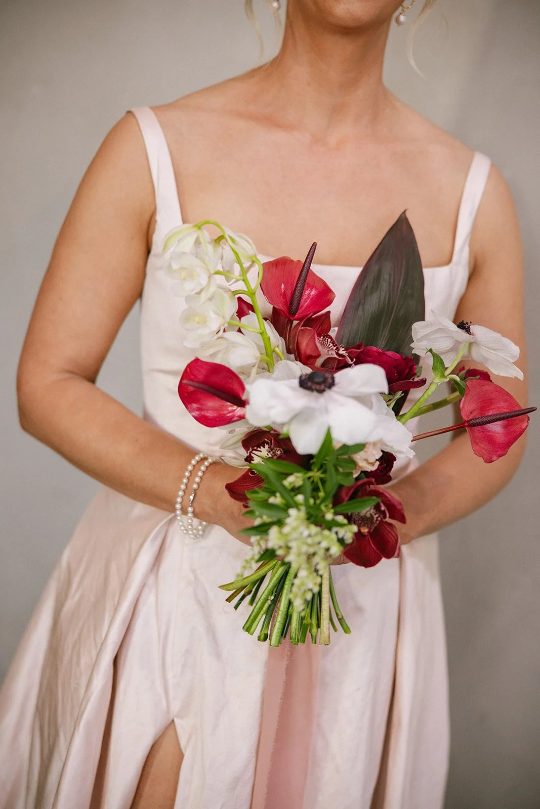 A woman in a light-colored dress holding a bouquet of red, white, and green flowers, wearing pearl bracelets and earrings.