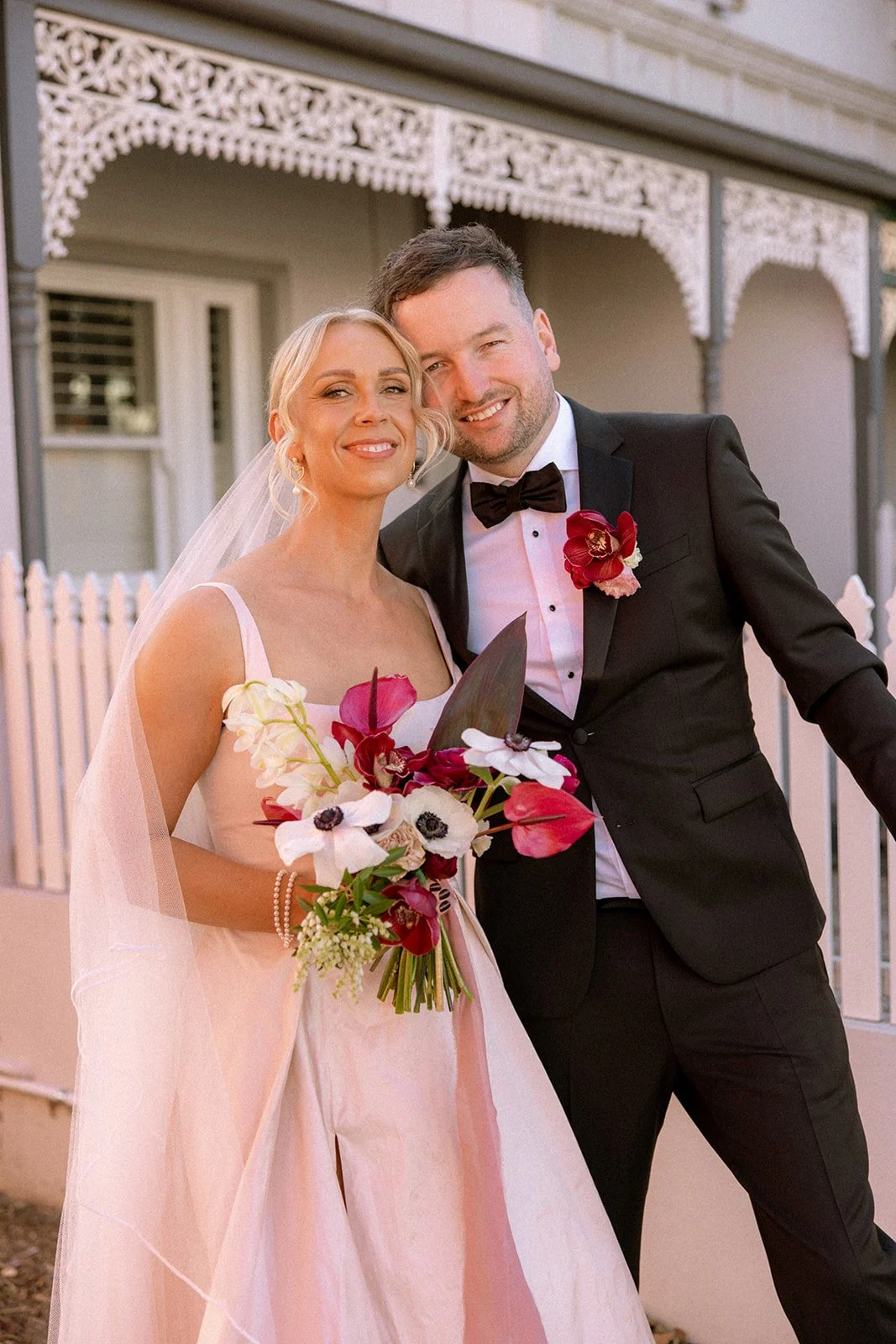 A bride and groom smile and pose together, the bride holding a bouquet of colorful flowers and the groom wearing a black suit with a red flower boutonniere, standing outside a house with decorative trim.