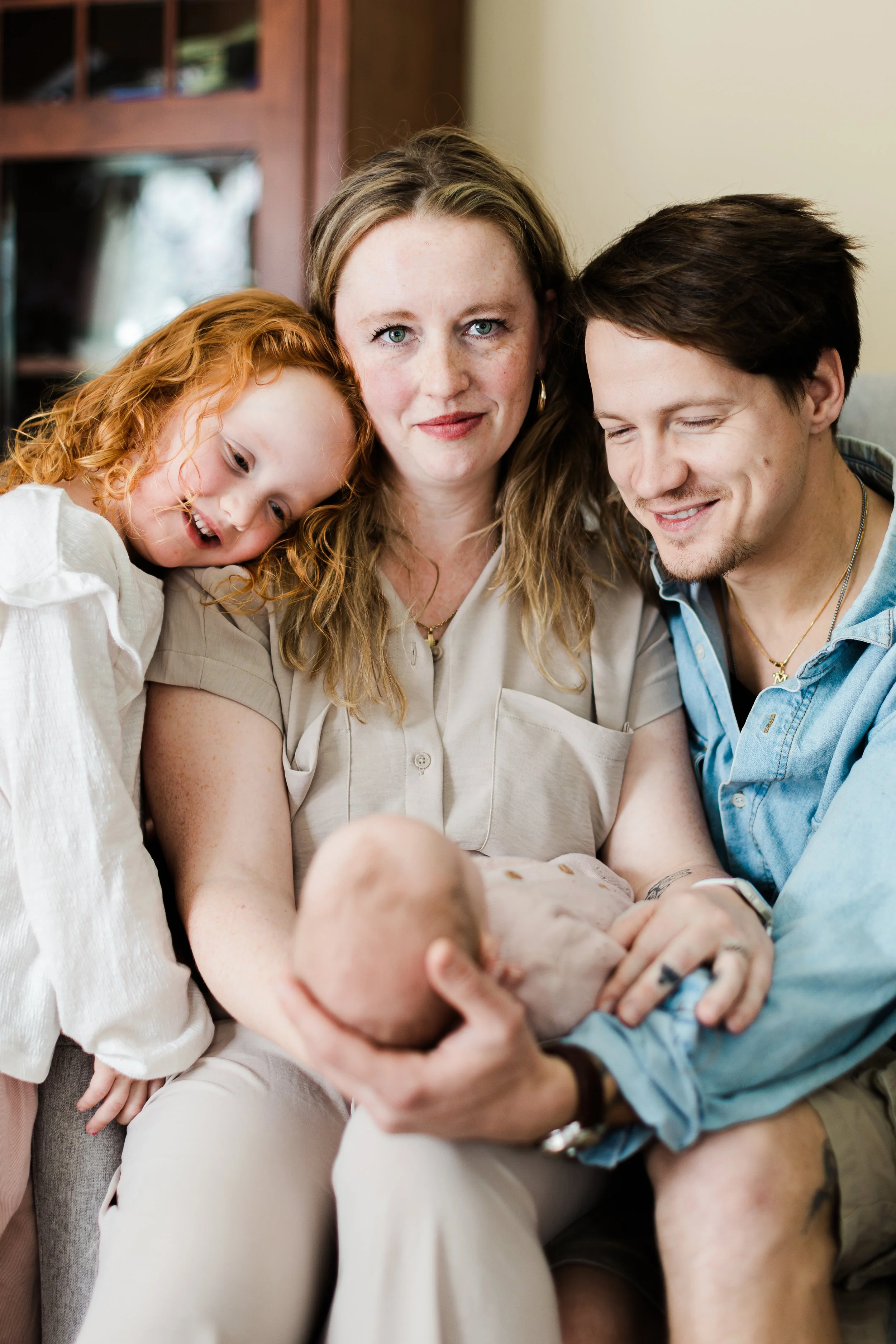 A woman, a man, a young girl with red curly hair, and a newborn baby sitting together indoors, holding the baby, with warm smiles on their faces.