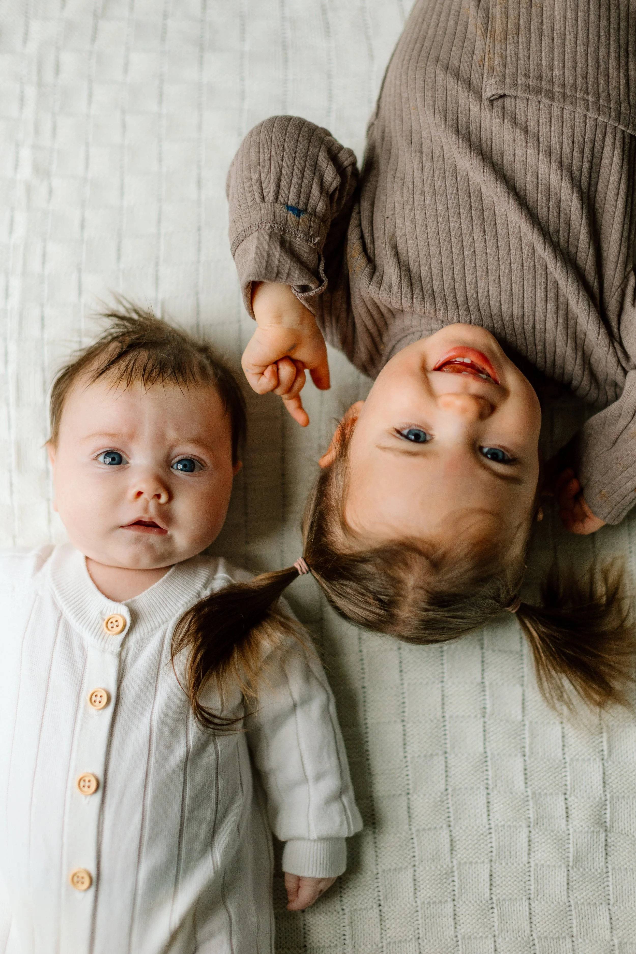 Two young girls lying on a cream-colored textured blanket, one smiling and the other looking at the camera with a serious expression.
