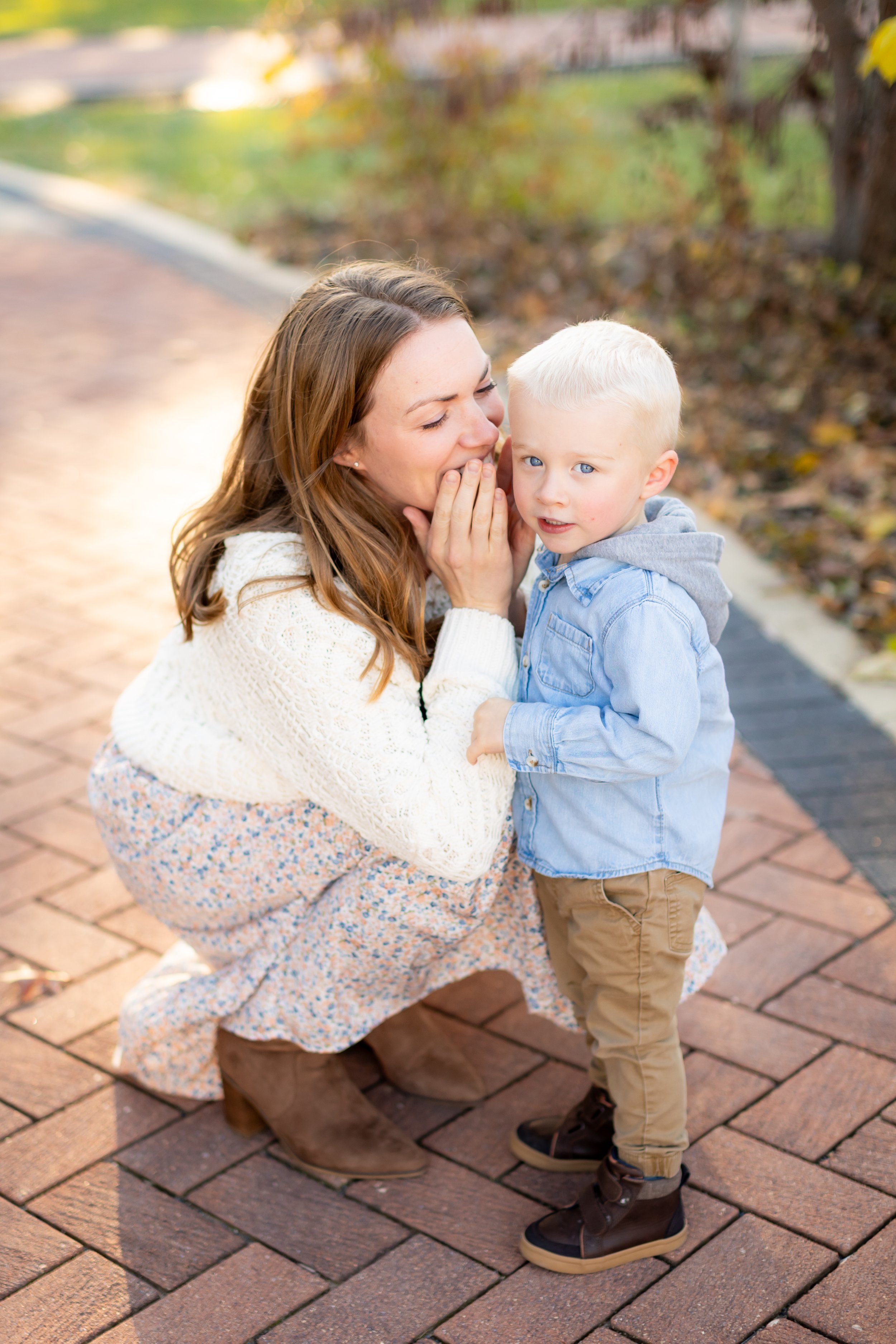 A woman squatting on a brick sidewalk, whispering to a young blonde boy with blue eyes, while the boy looks at the camera surrounded by autumn leaves and trees.