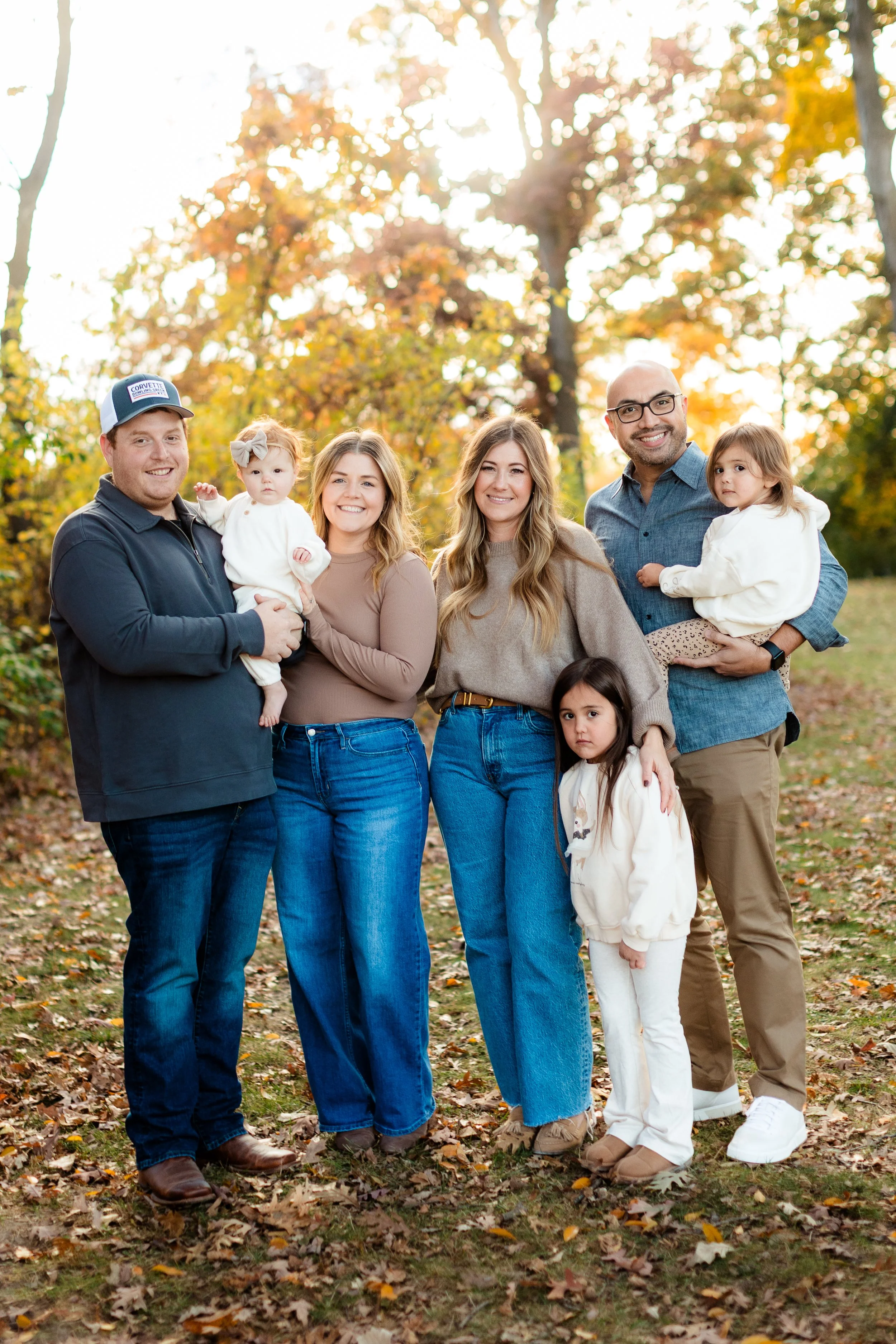 Family portrait outdoors in autumn with trees and fallen leaves, six people including two young children, smiling and posing for the photo.