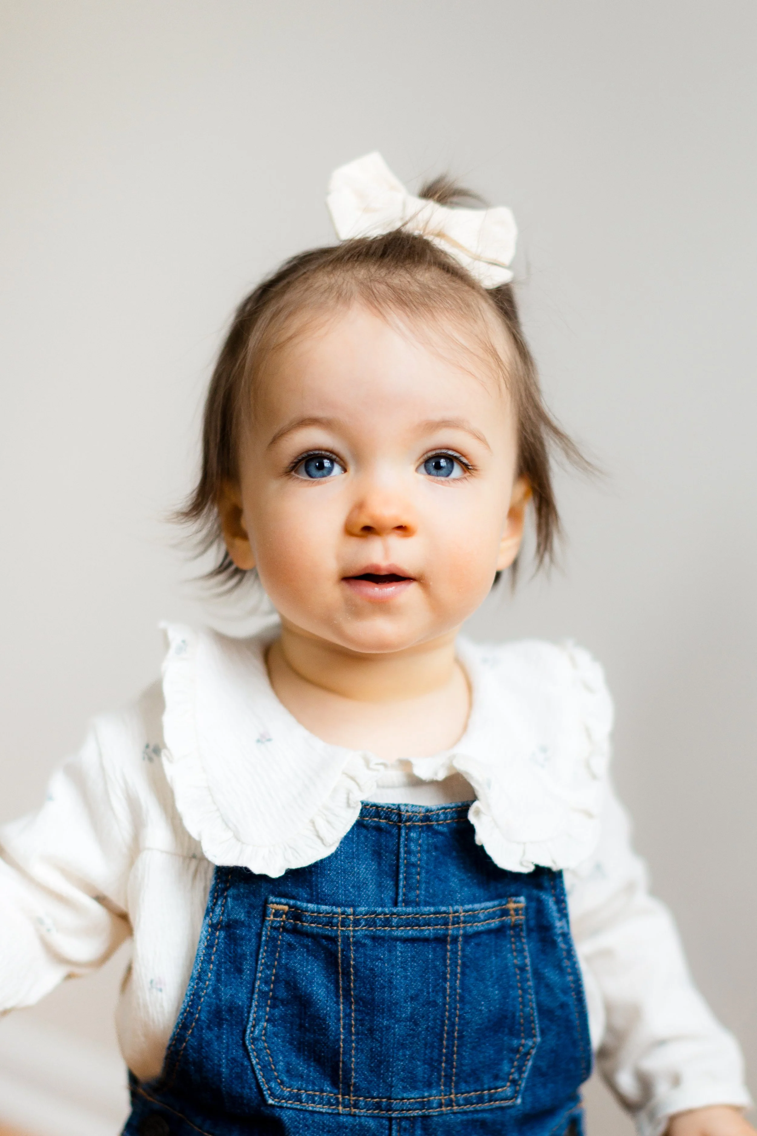 A young girl with blue eyes and light brown hair tied with a white bow, wearing a white blouse with ruffled collar and blue denim overalls.