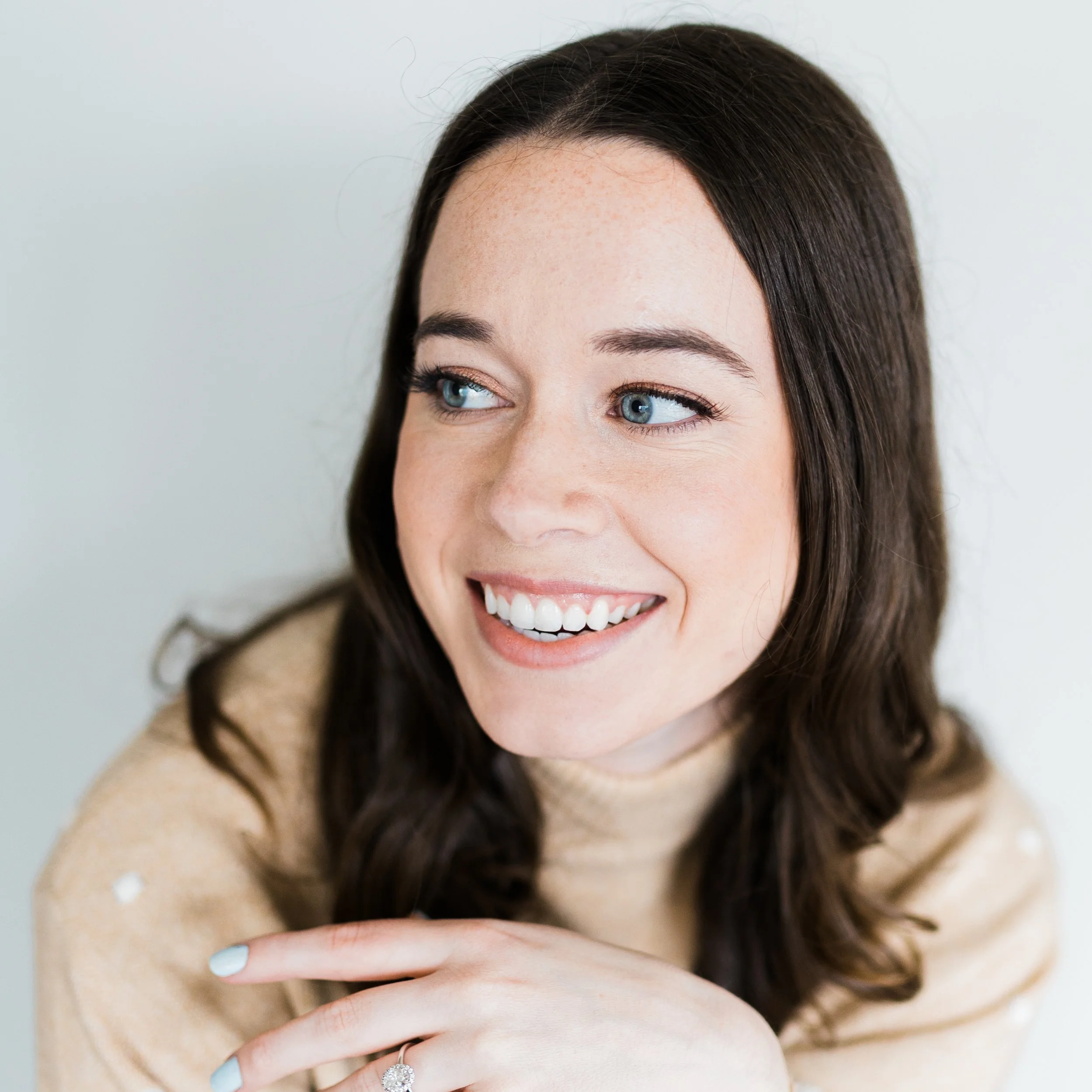 Close-up of a smiling woman with long brown hair and blue eyes wearing a beige sweater with small white dots, resting her hand on her shoulder with a ring visible.