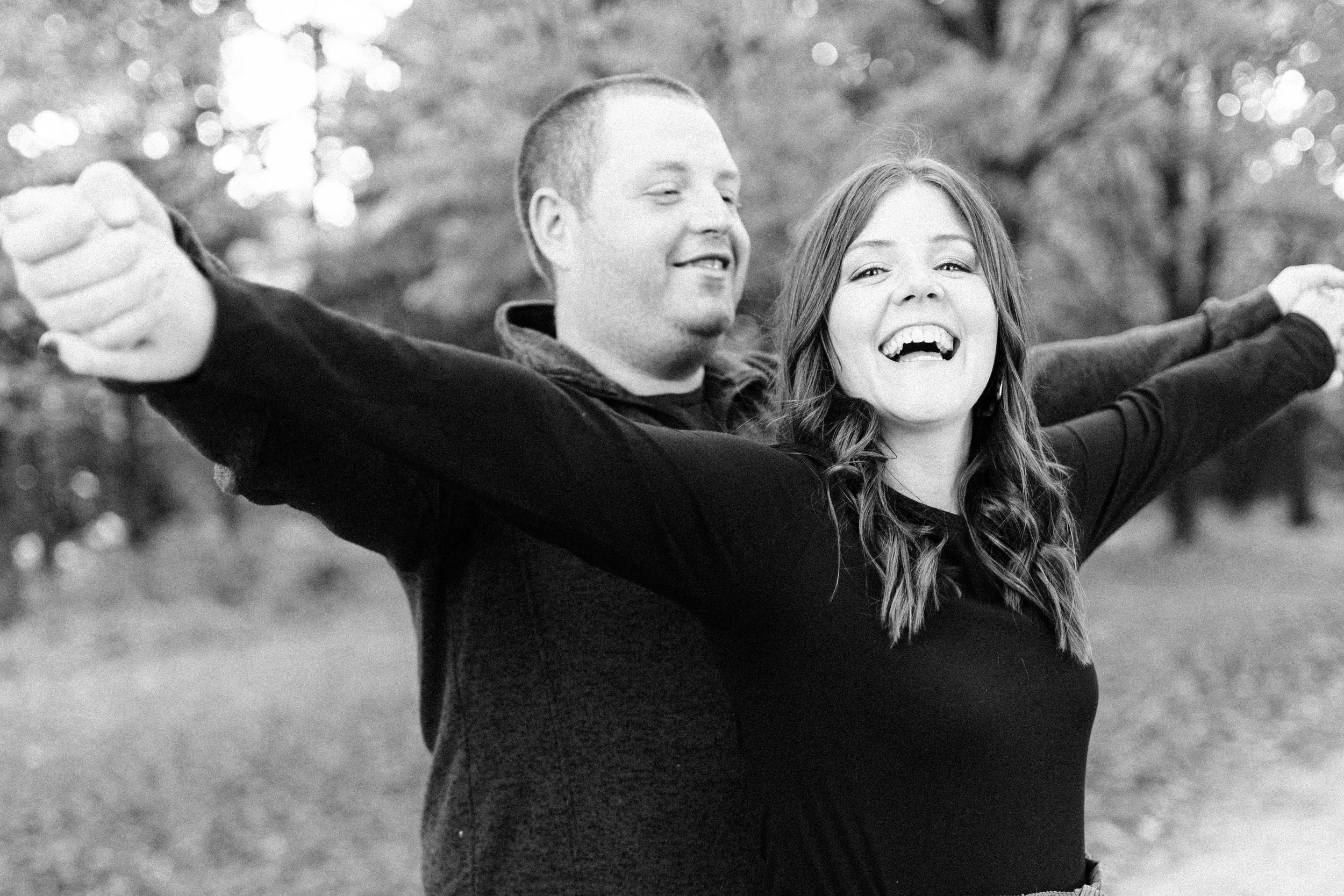 A smiling couple standing outdoors with arms outstretched, enjoying a sunny day in a park or natural setting.