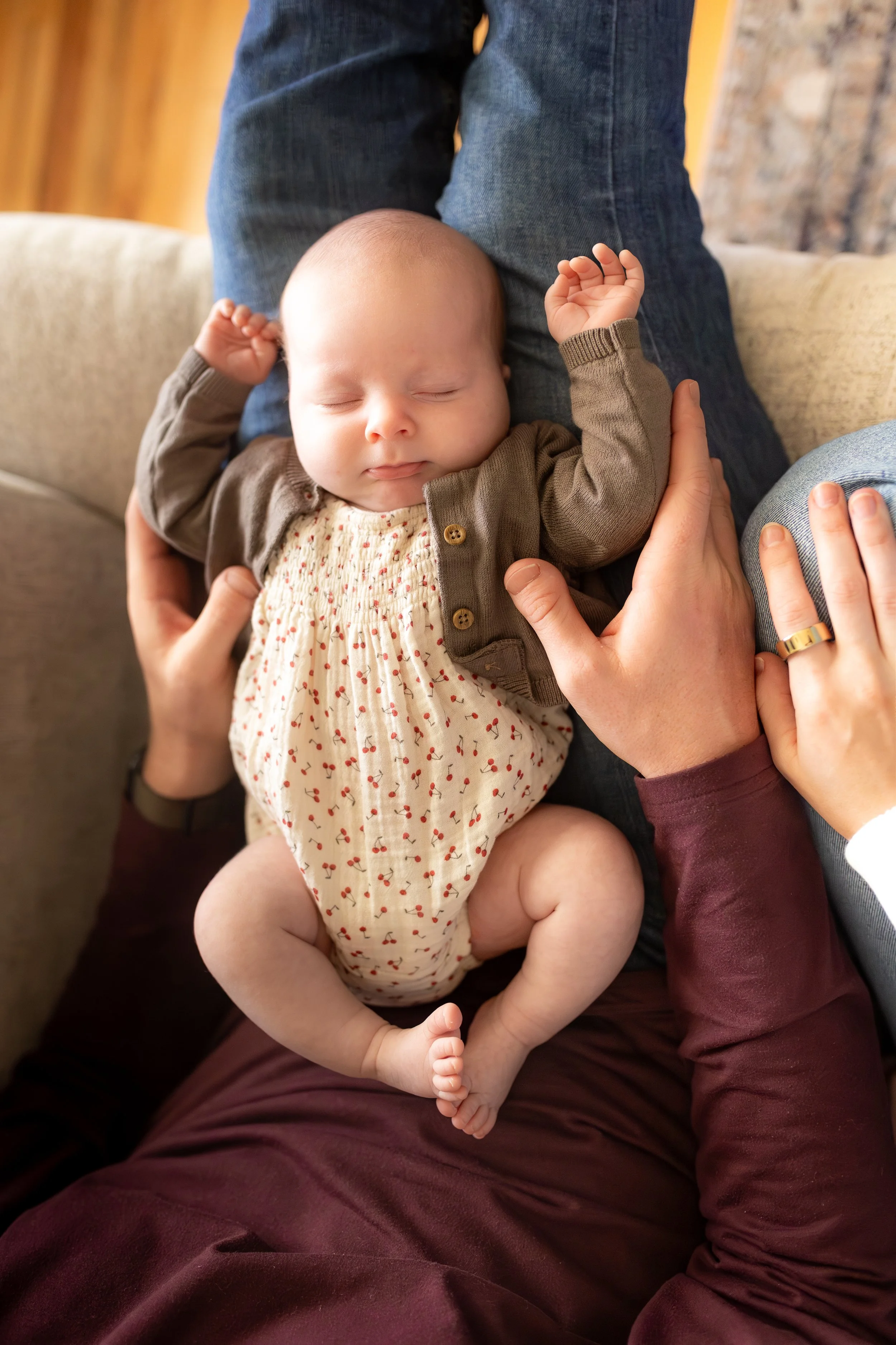 A baby sleeping peacefully on an adult's lap, with hands gently holding the baby's shoulders and arm, indoors with a beige sofa and wooden floor visible.
