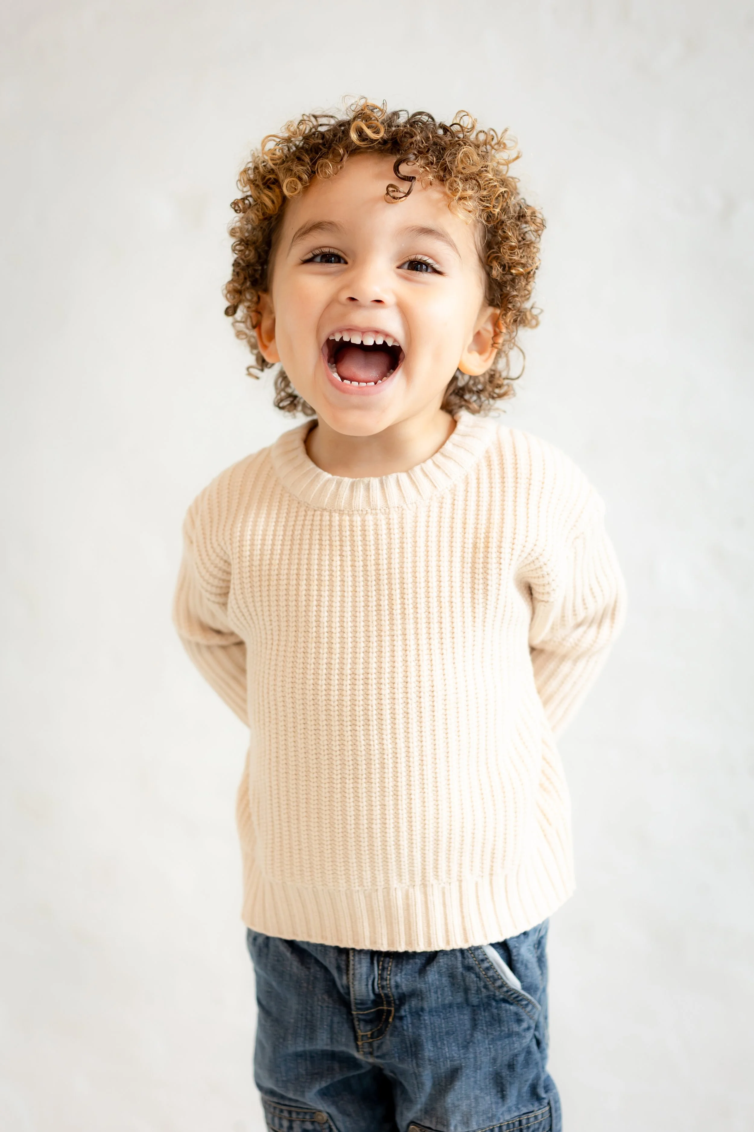 A young boy with curly hair smiling widely and laughing, standing against a plain light-colored background.