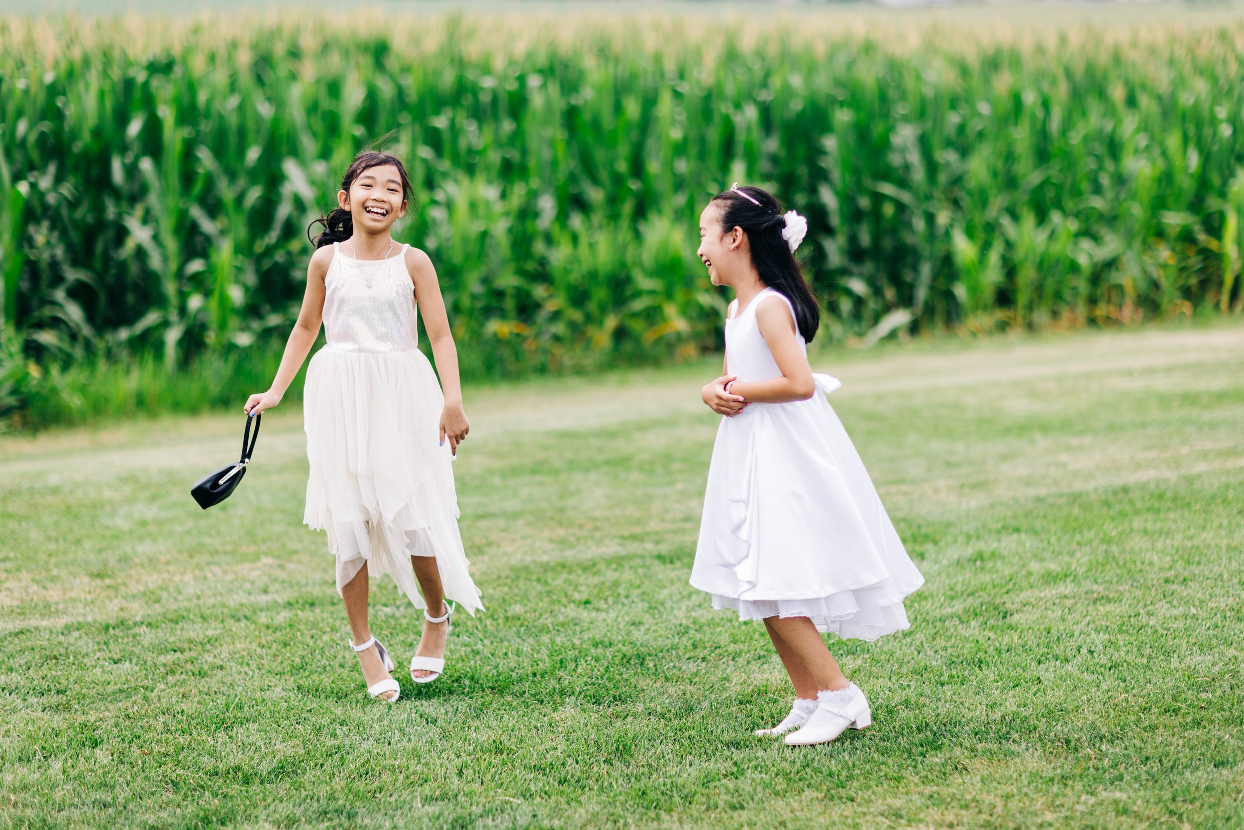 Two young girls dressed in white dresses, laughing and playing on a grassy field with a cornfield in the background.