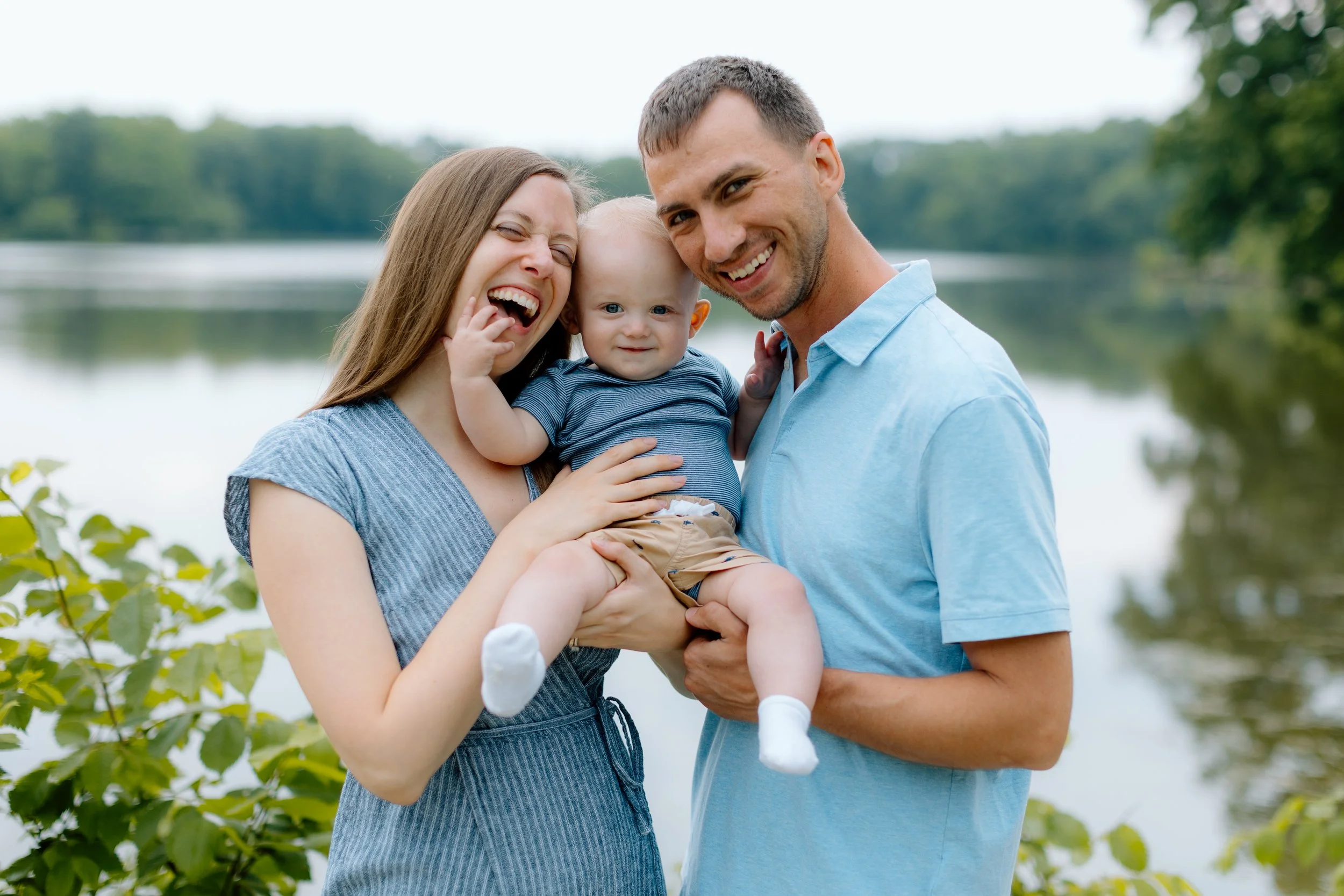 A happy family of three standing outdoors near a lake, with the mother laughing, the father smiling, and their baby grandson looking at the camera