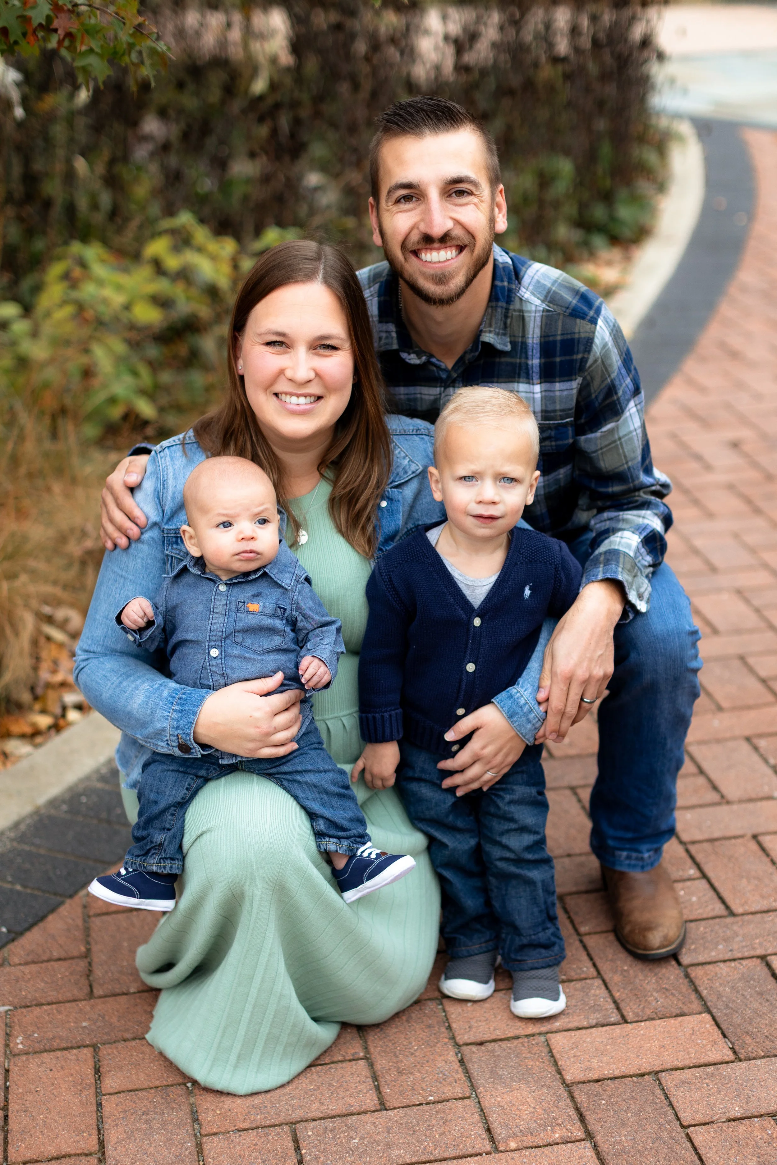 A smiling family of four posing outdoors on a brick walkway during fall. The mother, kneeling, holds a toddler with a serious expression. The father, kneeling, has his arm around the mother and is smiling. The older son stands in front of the father, looking at the camera with a neutral expression.