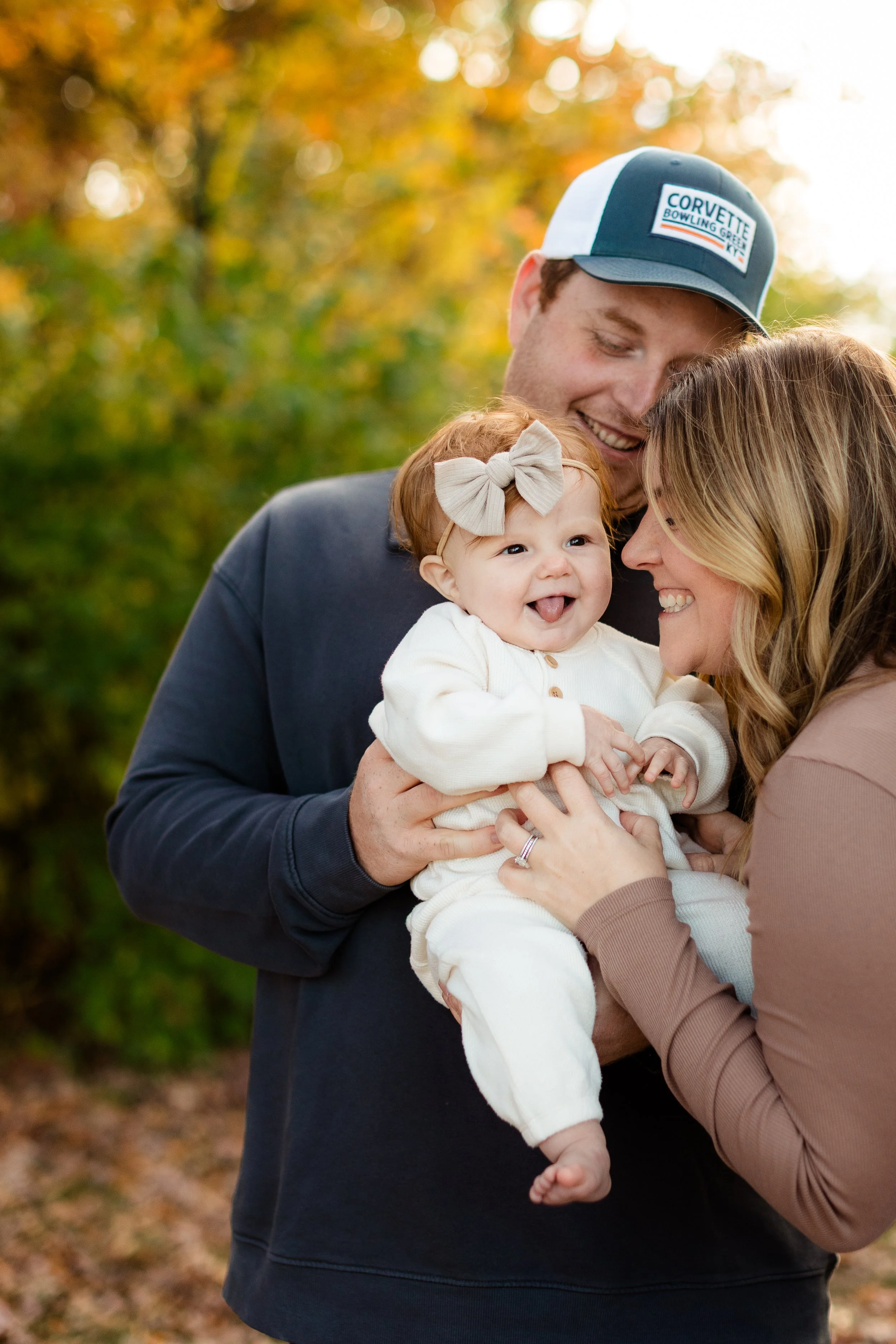 A family of three, including a man, woman, and baby, outdoors during fall, smiling and touching foreheads, with colorful autumn foliage in the background.