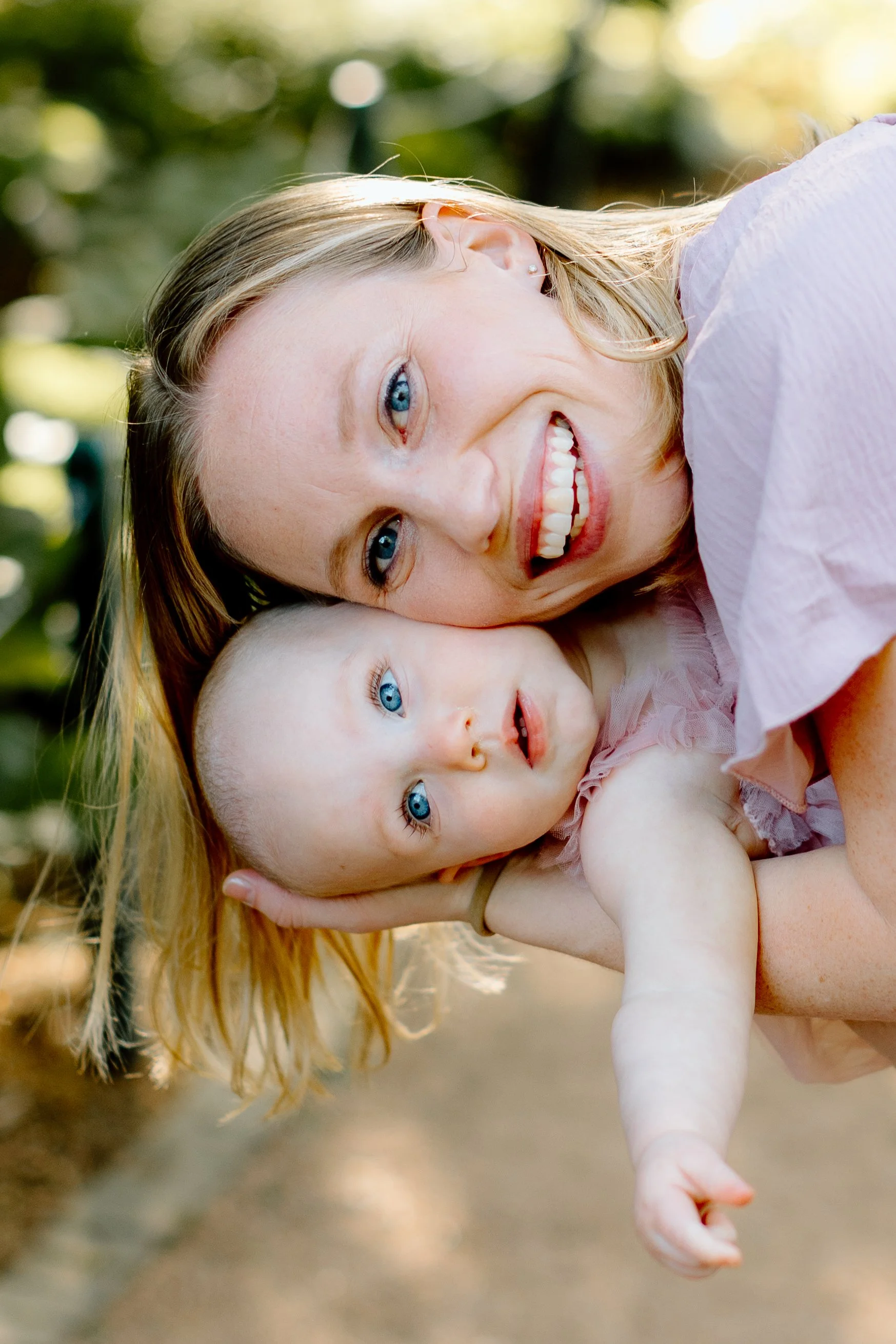 A woman and a baby lying close together outdoors, both with blue eyes and smiling, with the woman holding the baby's head.