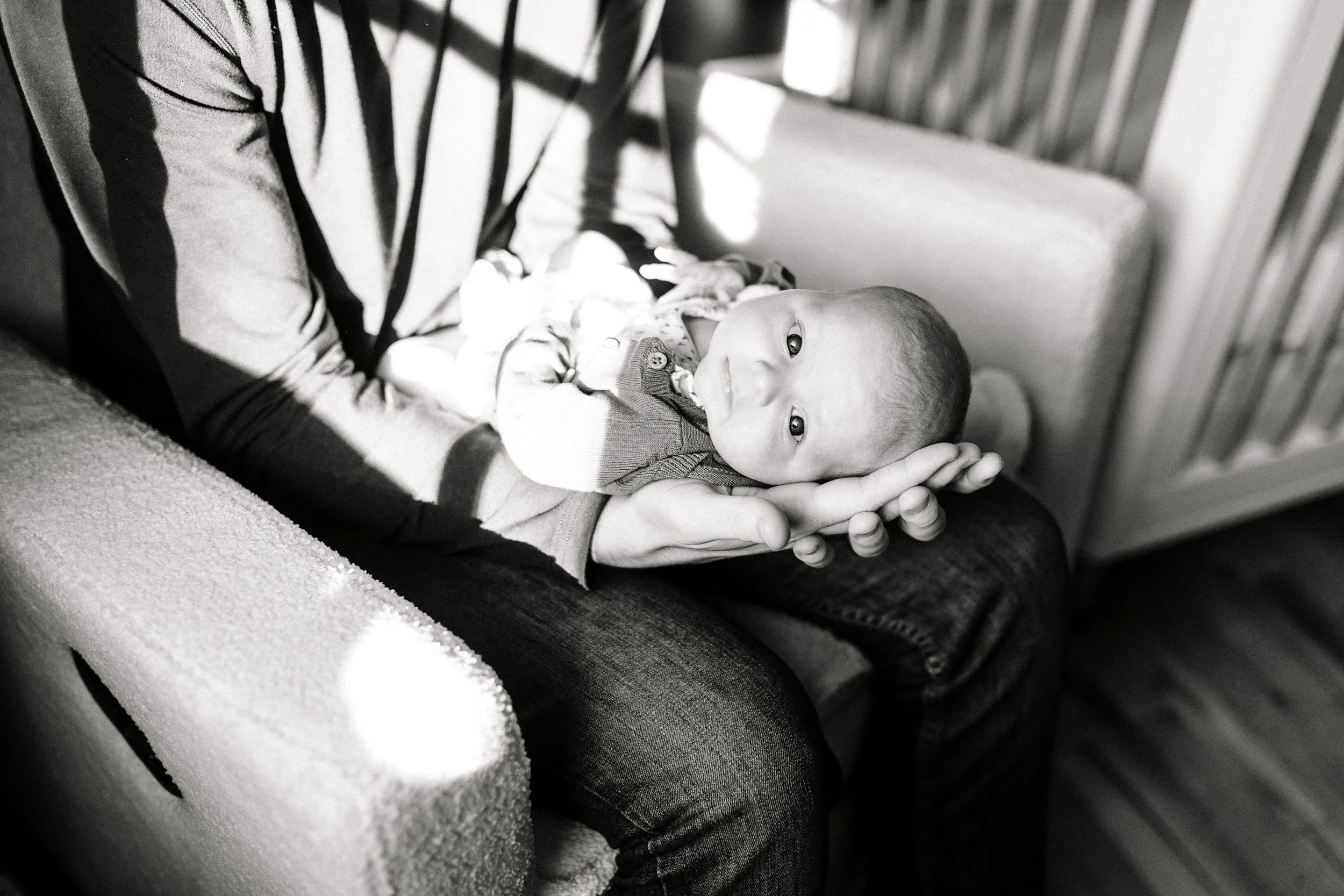 A baby lying in an adult's hands, looking at the camera, with an adult holding the baby's head while sitting on a chair.