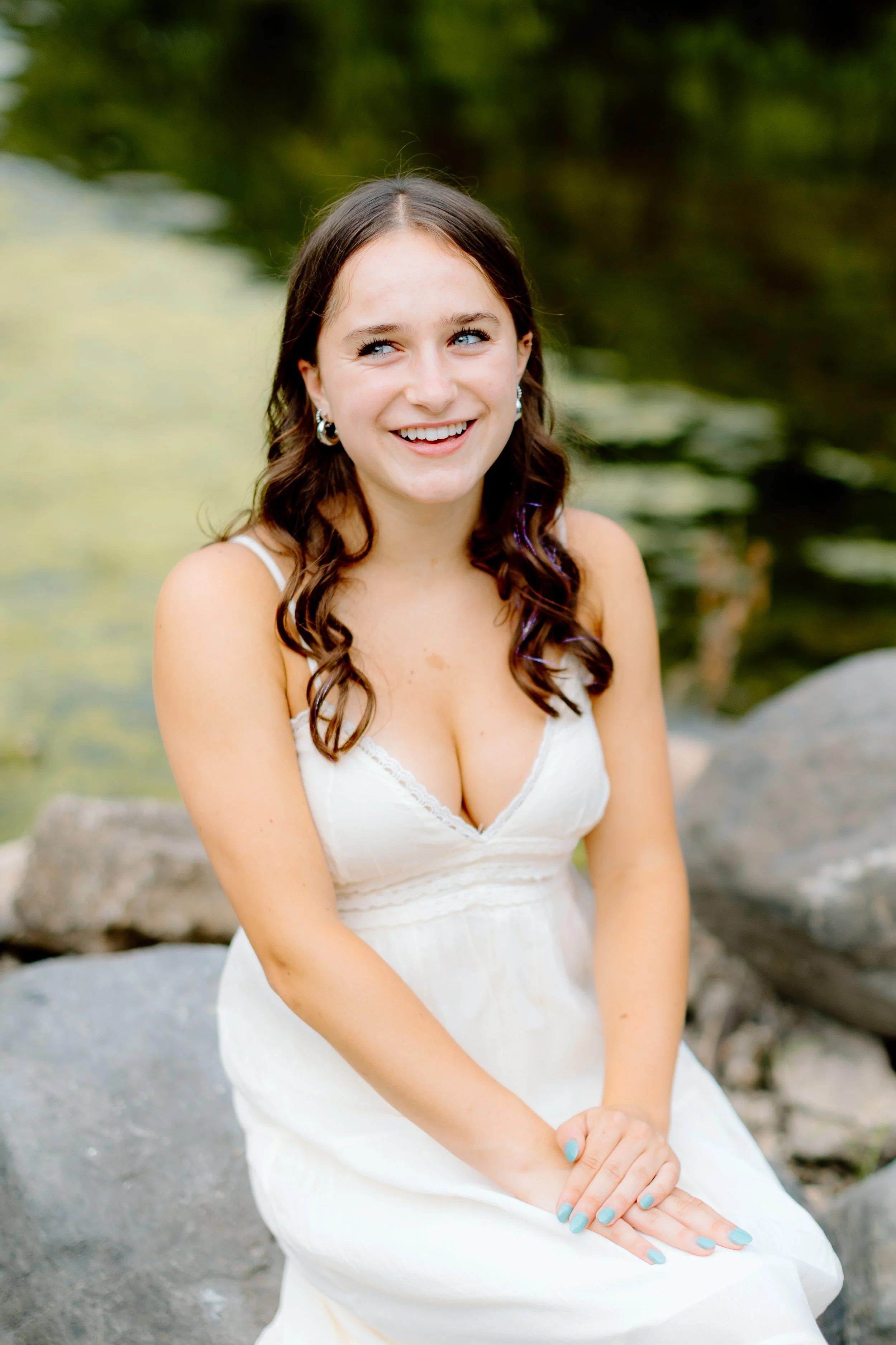 A young woman with long, wavy brown hair, wearing a white dress with thin straps, sitting on rocks near a body of water with greenery in the background, smiling and looking slightly to her right.