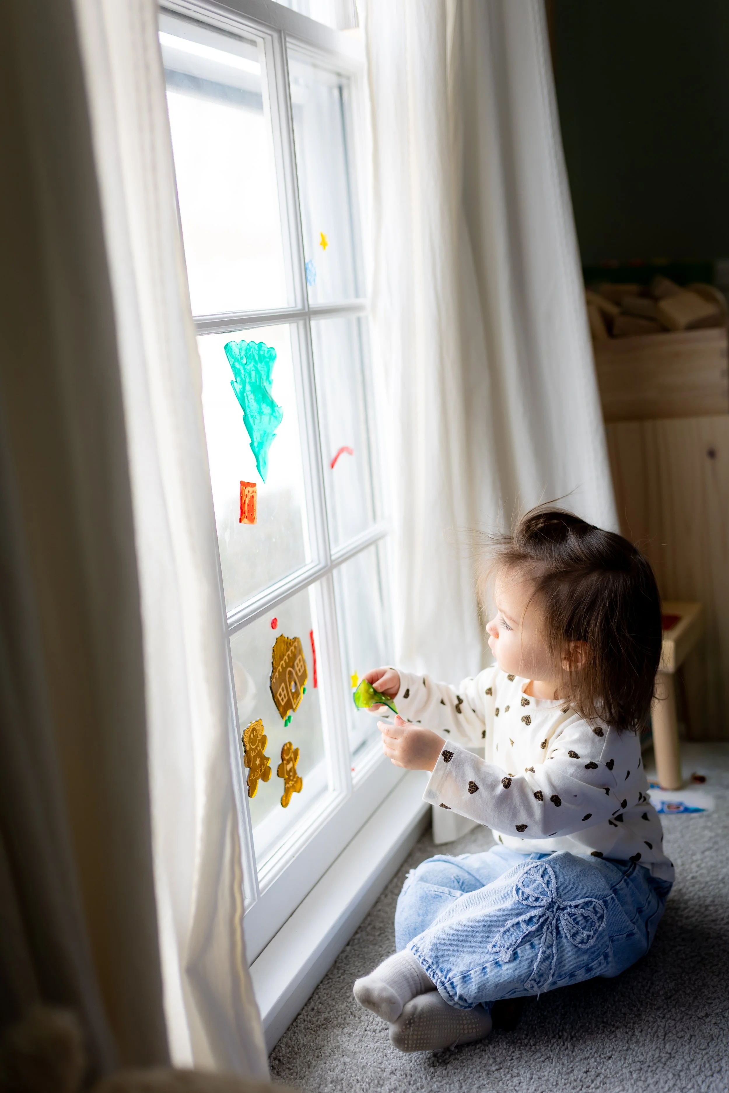 A young girl sitting on the carpeted floor by a window, decorating the glass with colorful stickers depicting a tree, gingerbread men, and houses.
