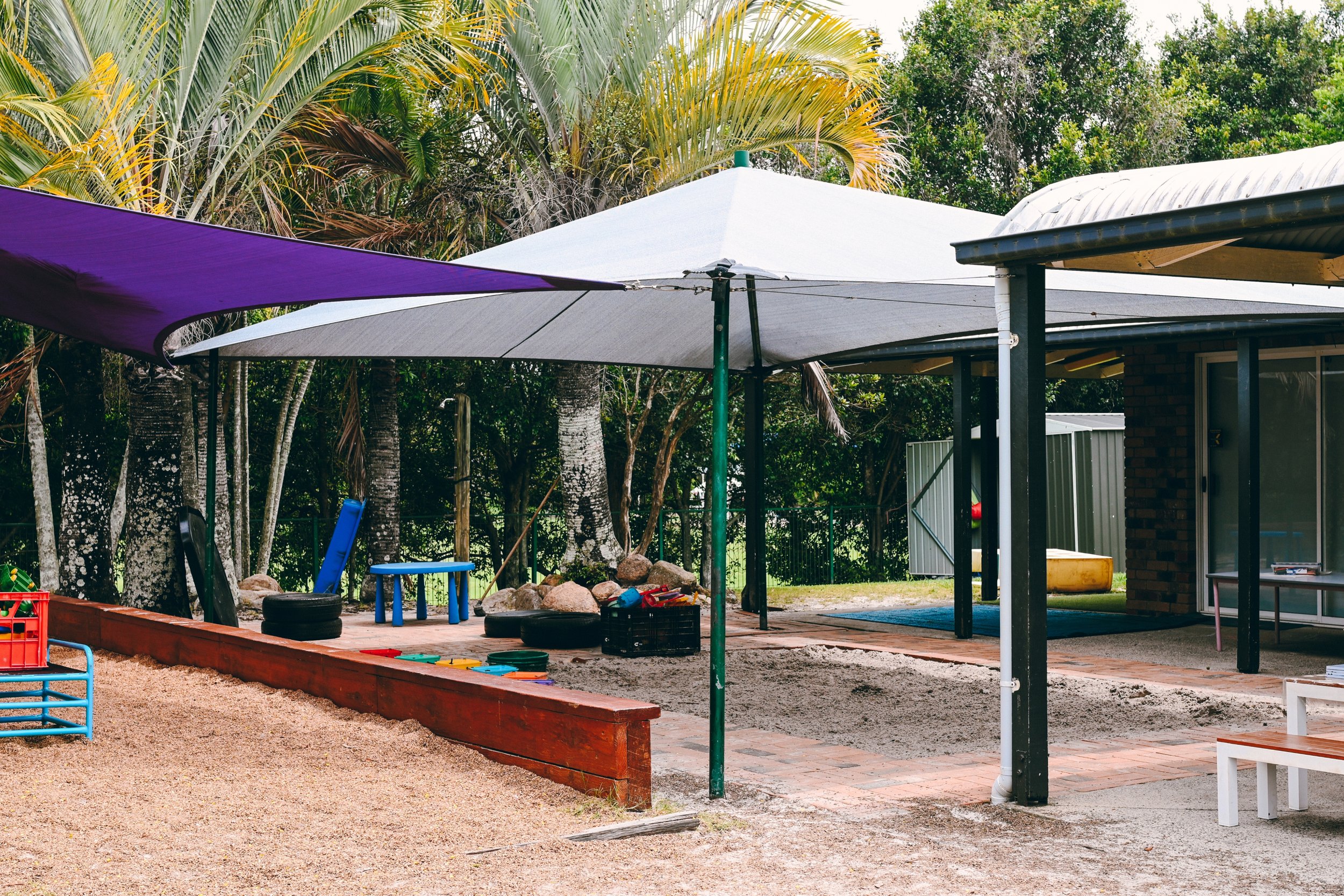 Outdoor playground area with shade sails, benches, and play equipment, surrounded by trees and foliage.
