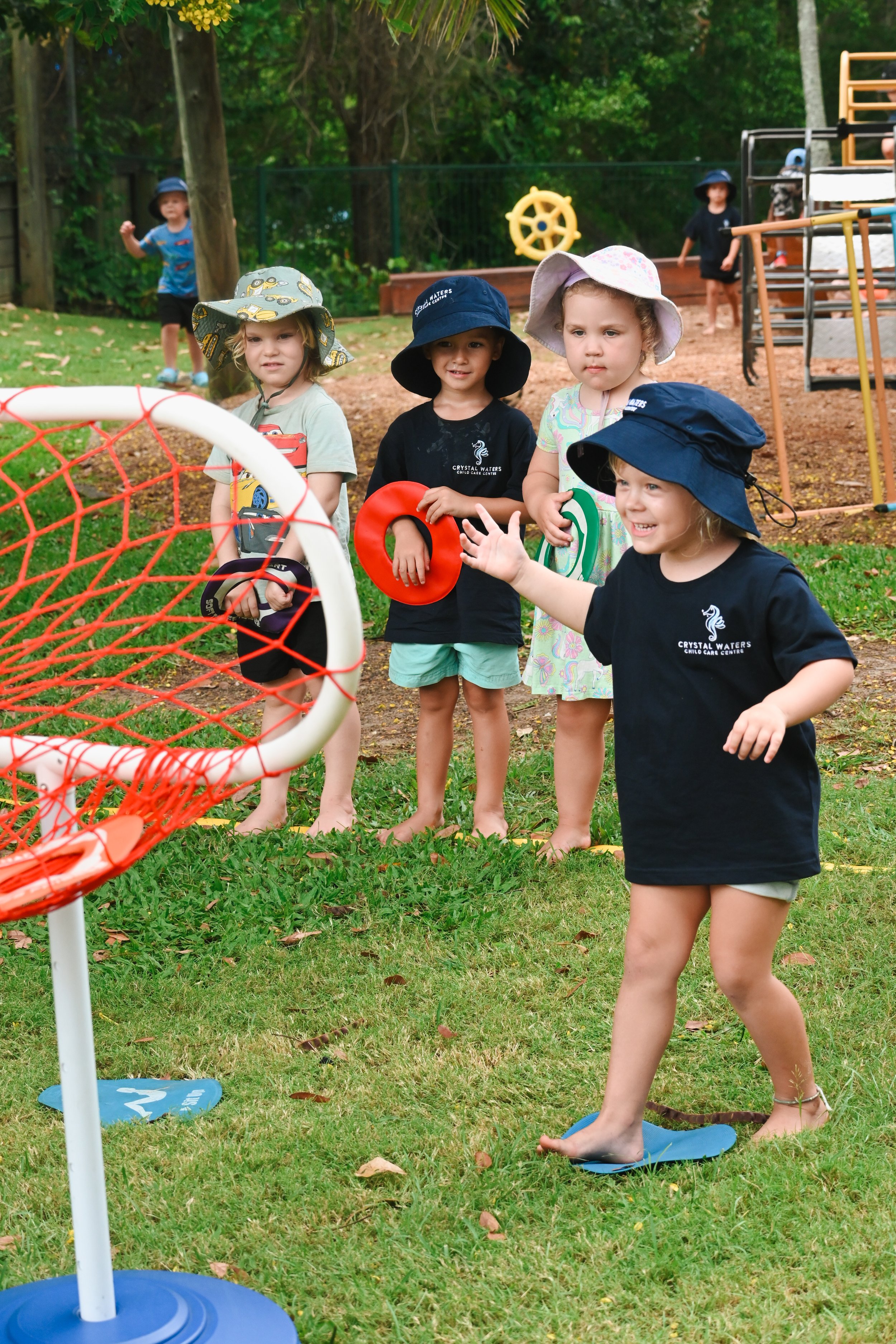 Children playing ring toss game in a grassy outdoor area, wearing sun hats and casual clothing, with trees and playground equipment in the background.