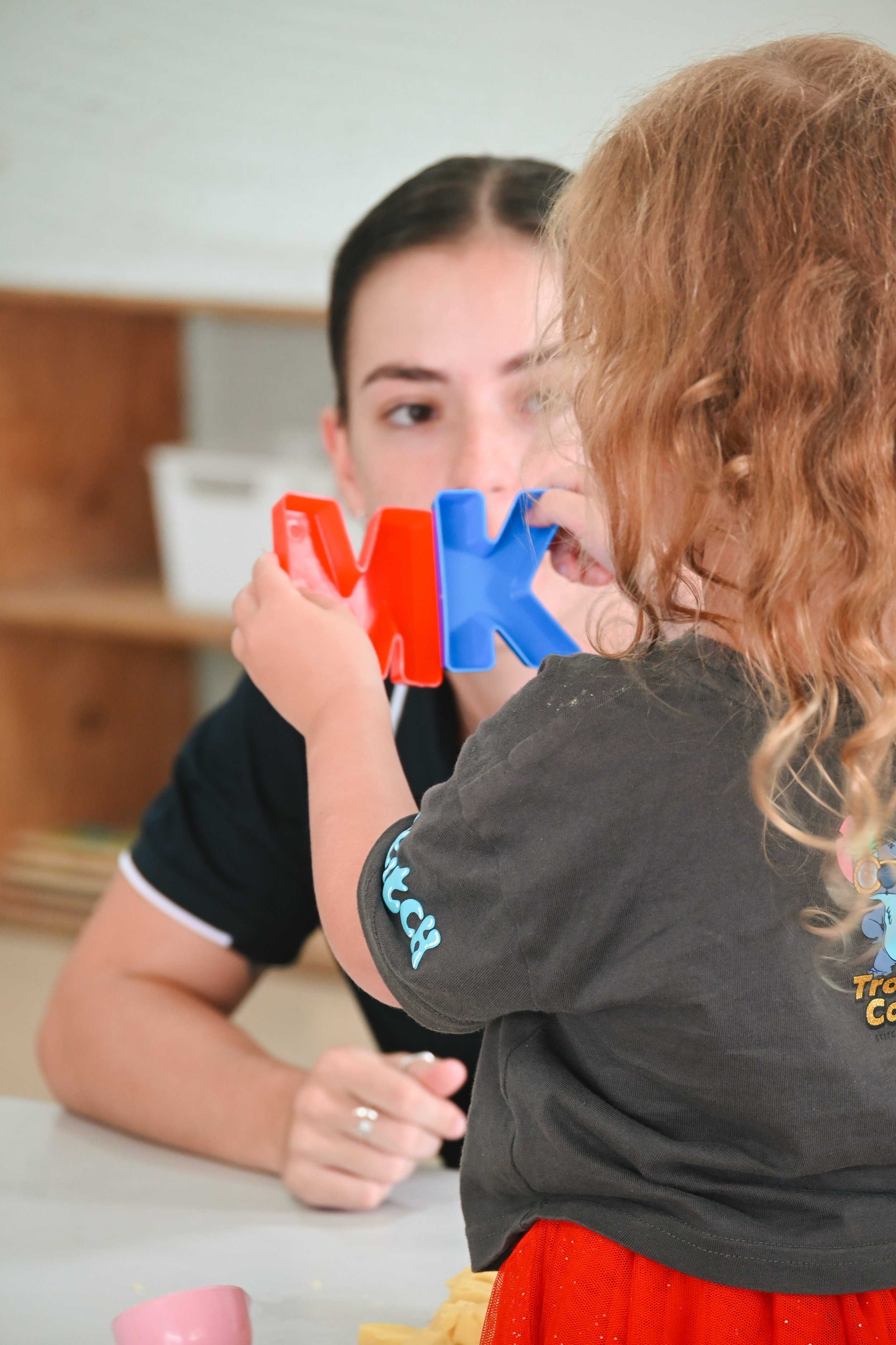 Child holds red and blue letters M and K with adult observing.