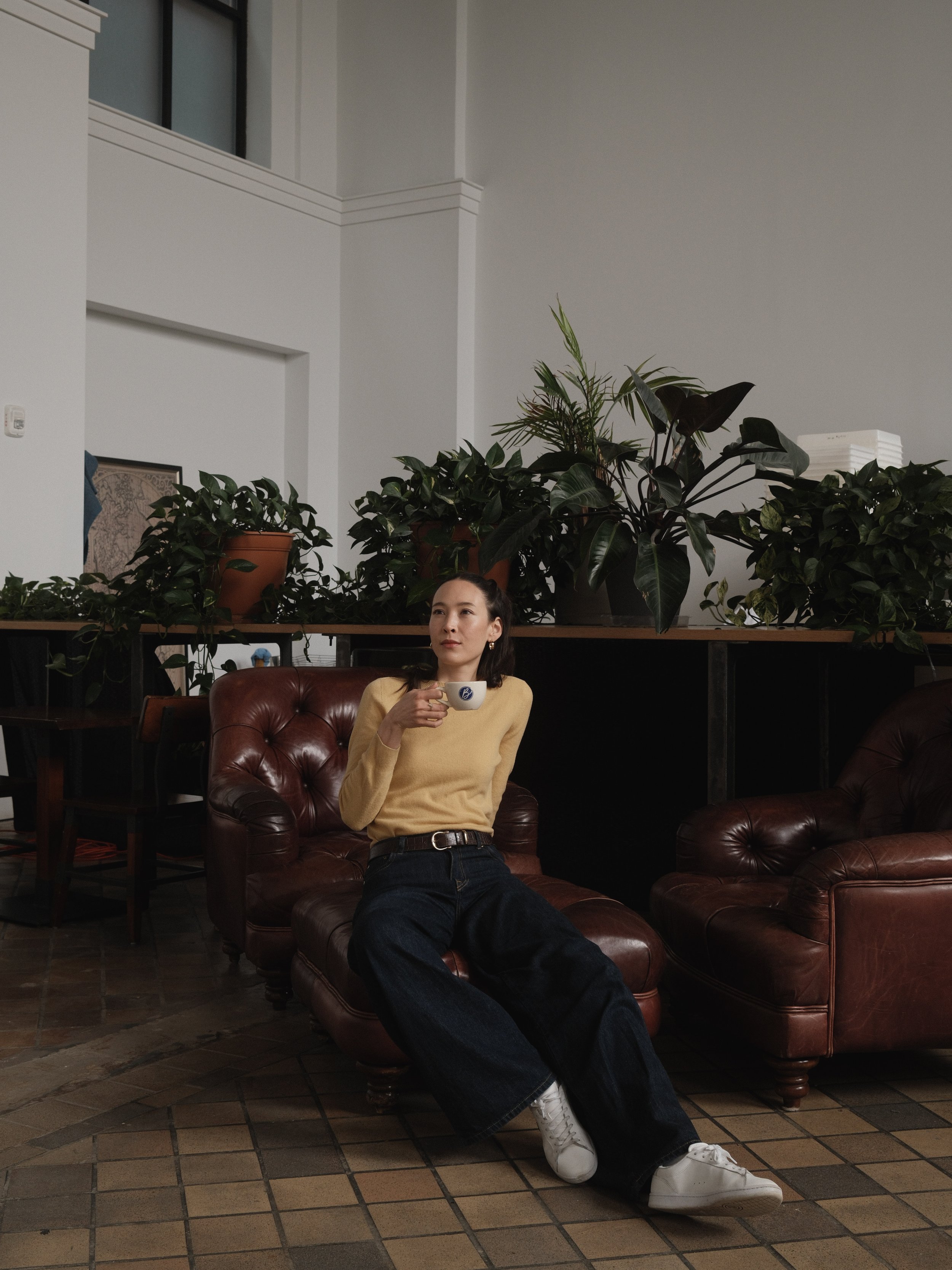 Tall Korean woman wears a yellow sweater tucked into long blue jeans, as she is seated on a dark brown leather couch.