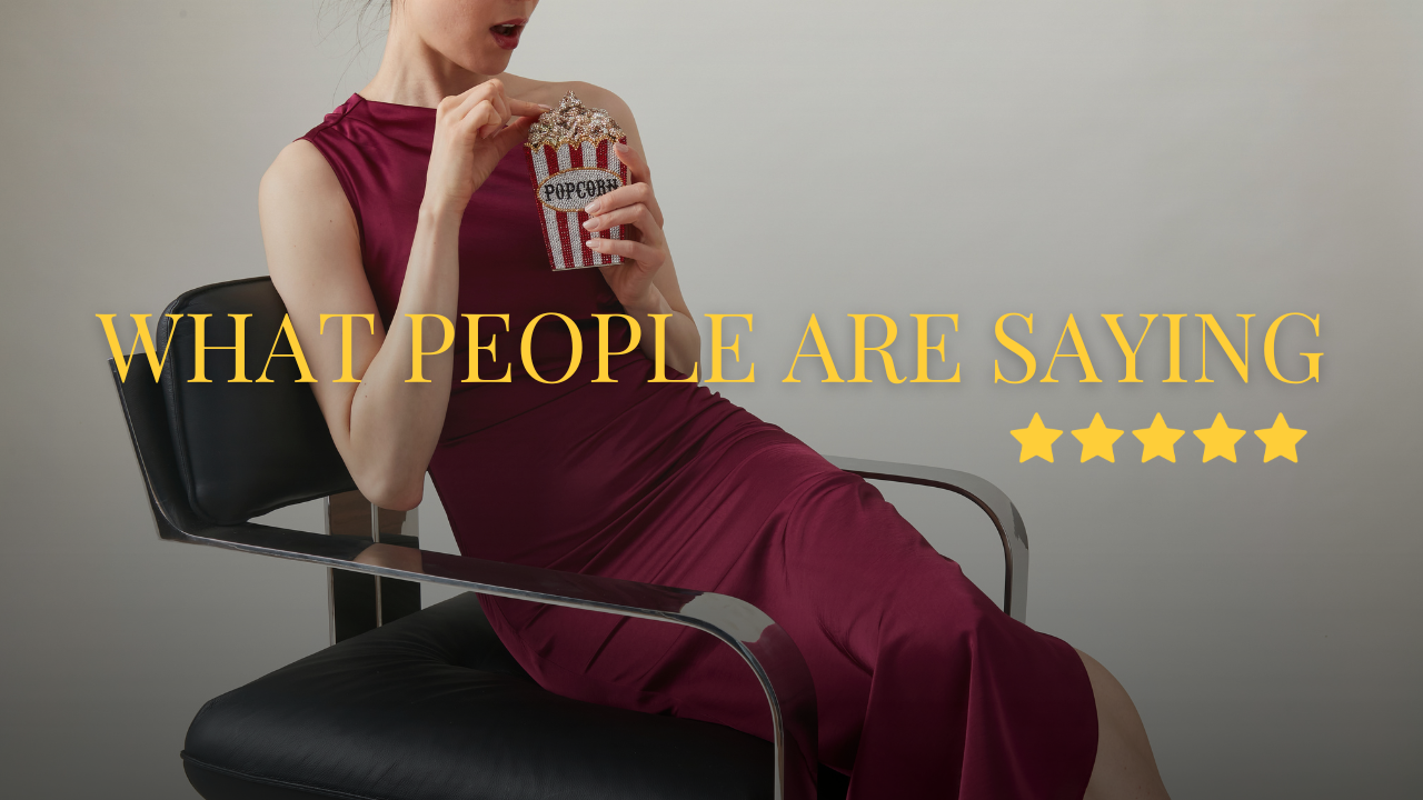 A beautiful, slim girl sits on a black chair, wearing a wine colored silk dress and holding a jeweled bag shaped like a popcorn box