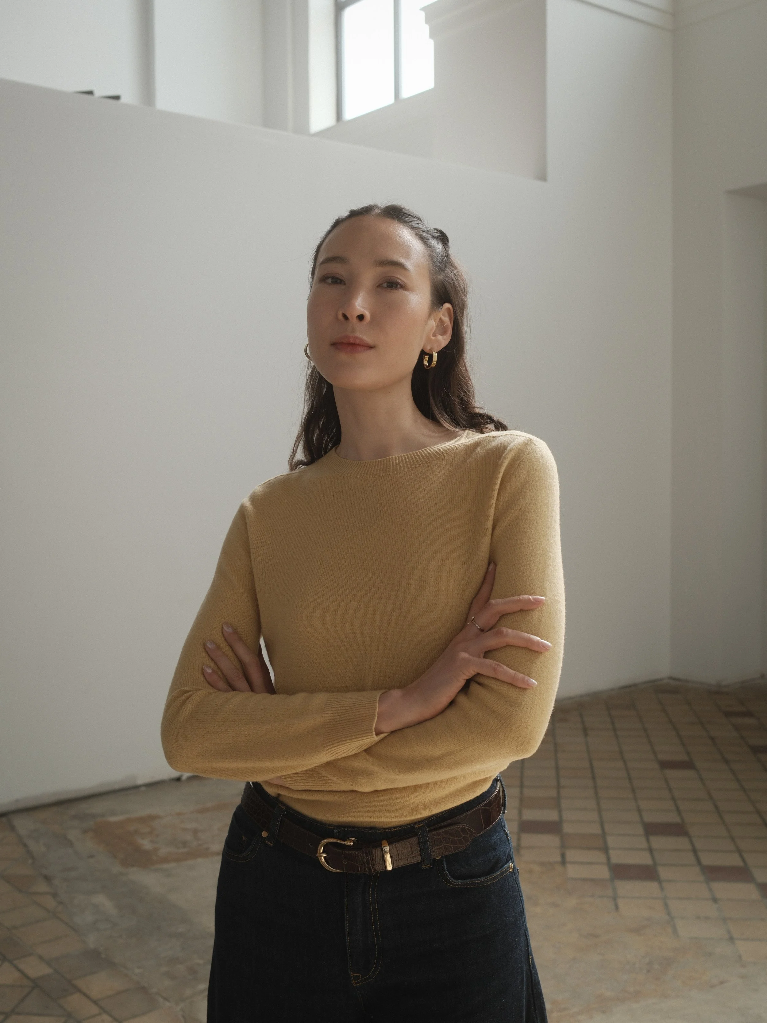 Korean model and actress Rebecca Rose wears a mustard yellow sweater and gold hoop earrings against a white wall in a sunny cafe