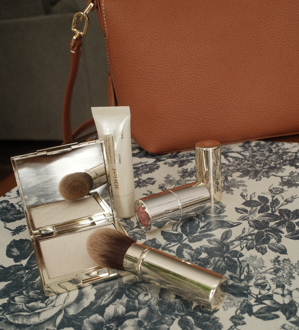 A lip stick, powder and brush from Jouer Cosmetics on a flower patterned table.
