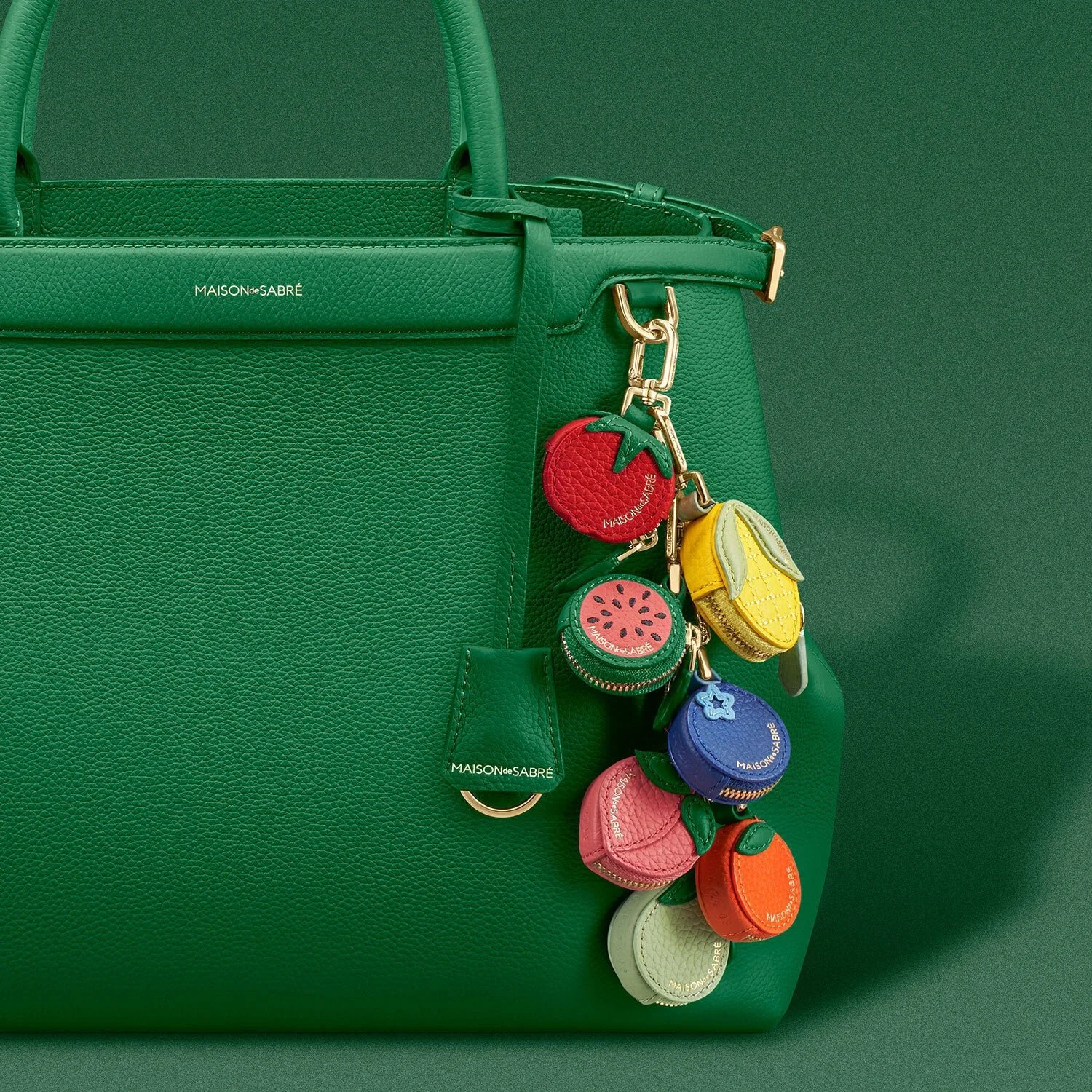 A green Maison de Sabre leather bag with fruit bag charms dangling off of the handle.h