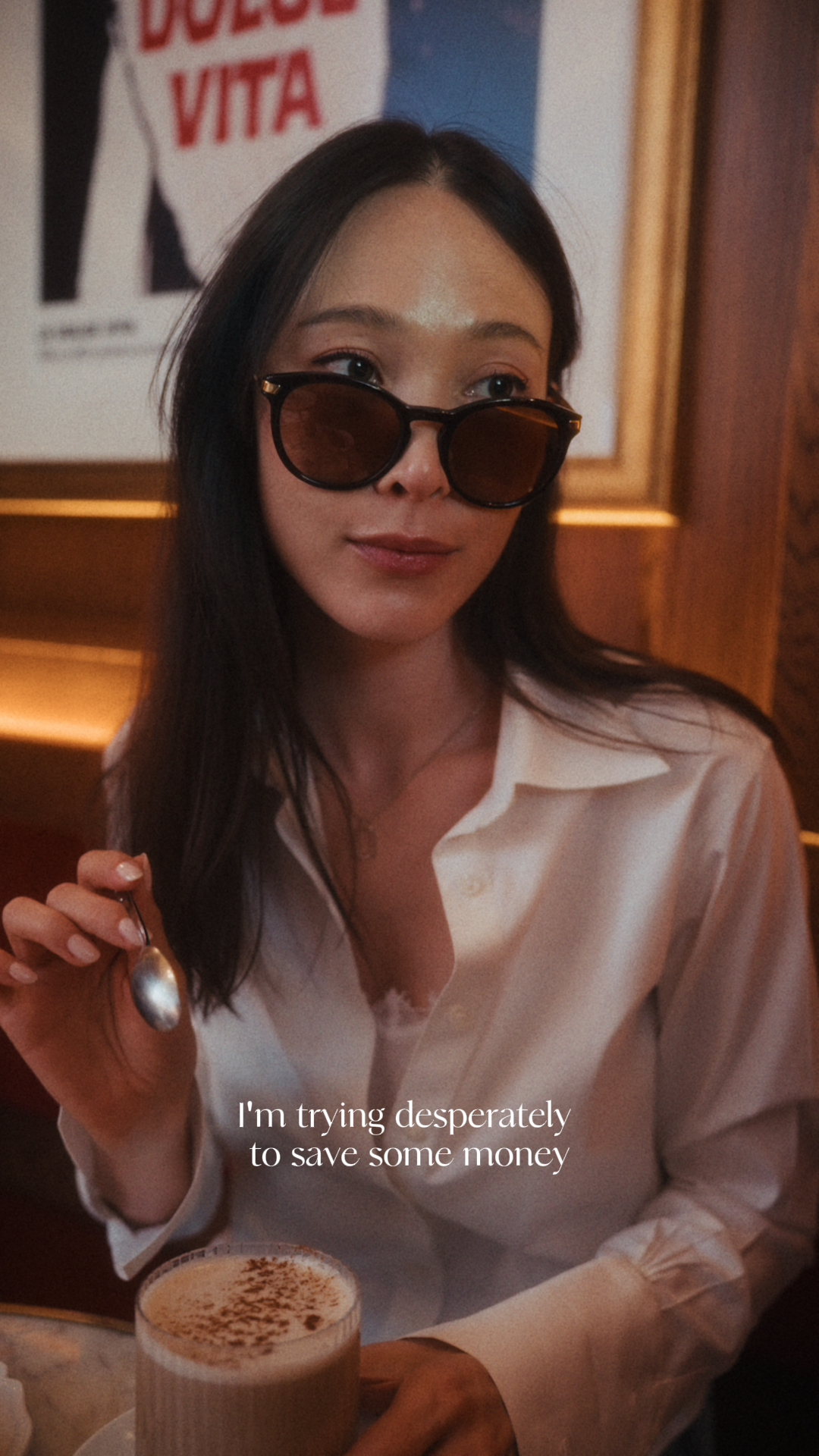 A beautiful girl wearing sunglasses and a white button up is sitting at a cafe drinking a fancy latte