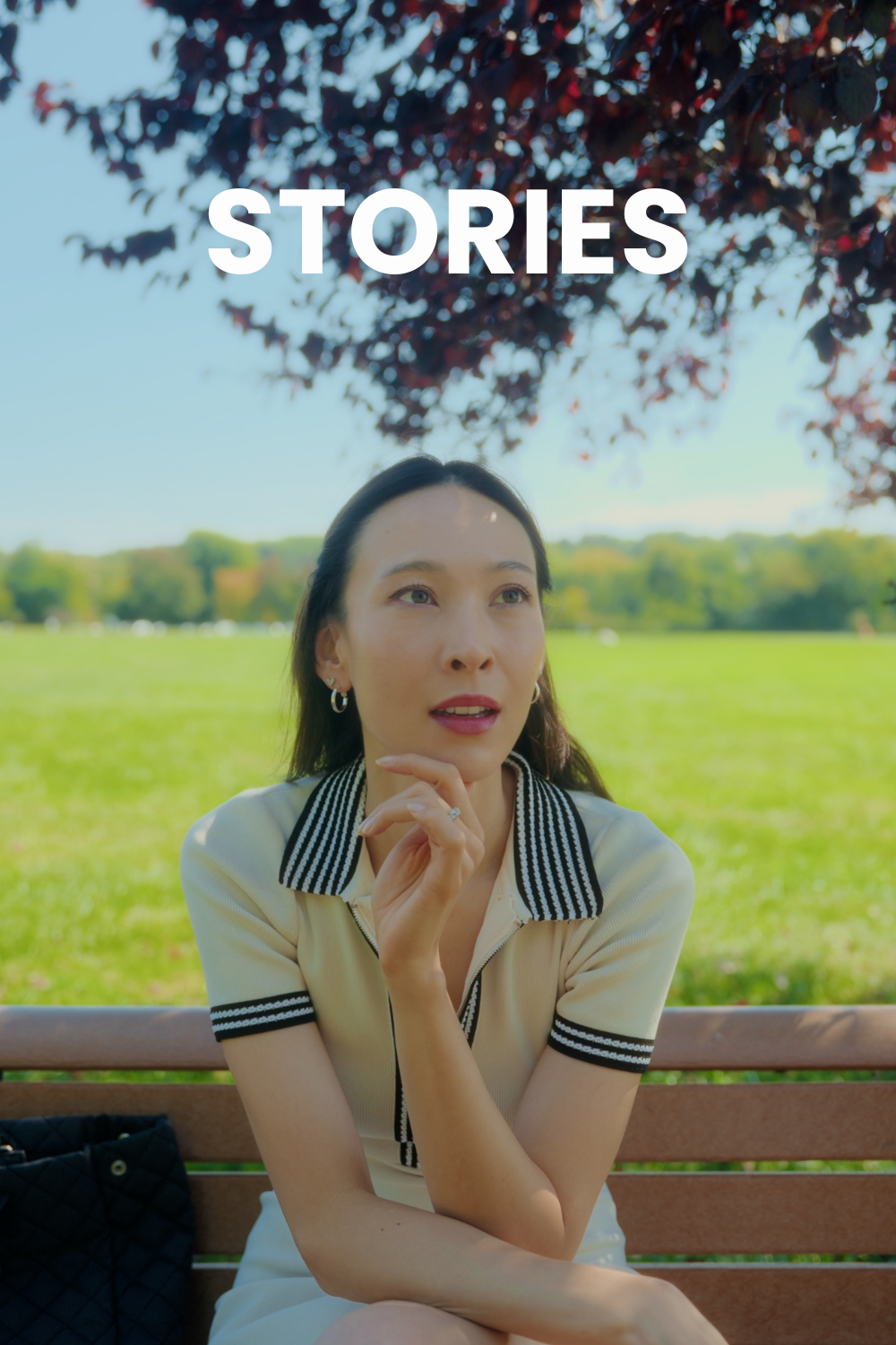 A Korean girl sits on a bench outside in a park, wearing a cream colored dress with a striped collar.
