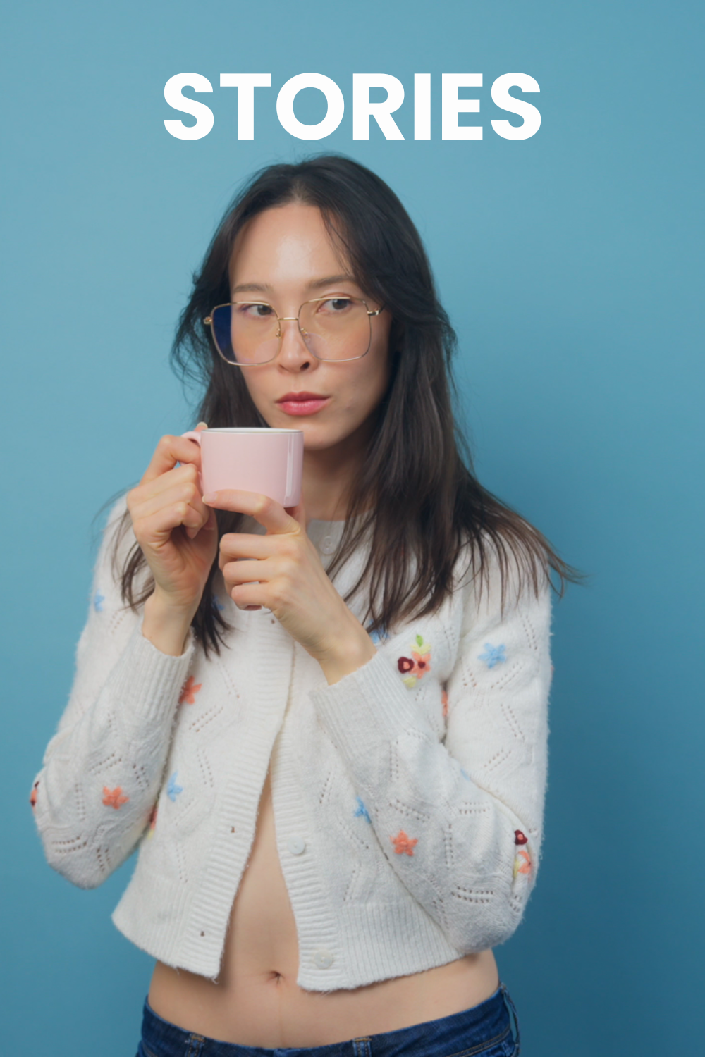 A beautiful Korean girl wearing a white sweater with flowers on it is holding a pink tea cup, is wearing gold rimmed glasses, and stands in front of a blue backdrop