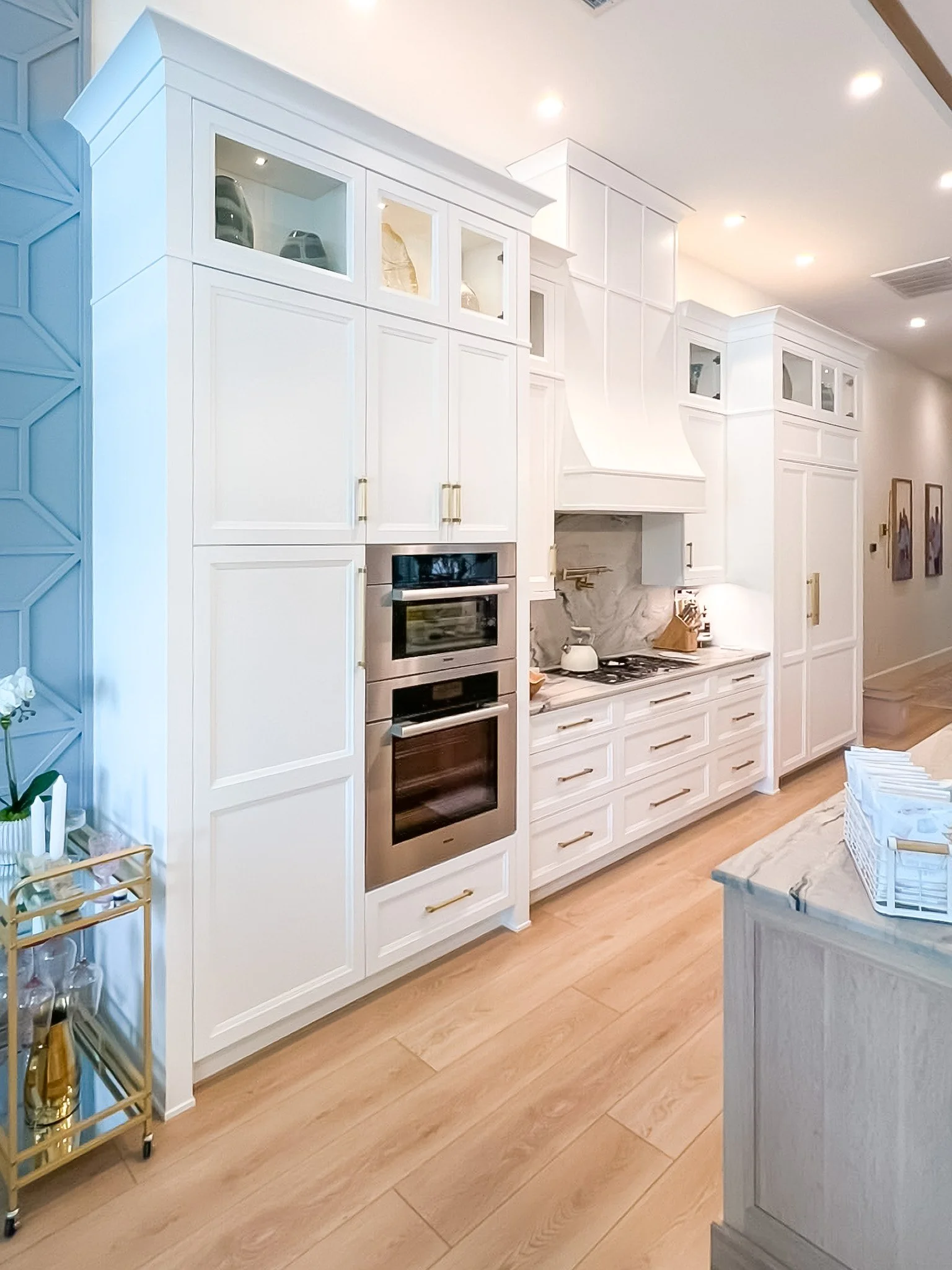 Modern kitchen with white cabinets, a kitchen island, built-in ovens, and a stainless steel range hood. Large windows with a view of lush greenery and a pink fence. Unique hanging light fixtures and a high ceiling.