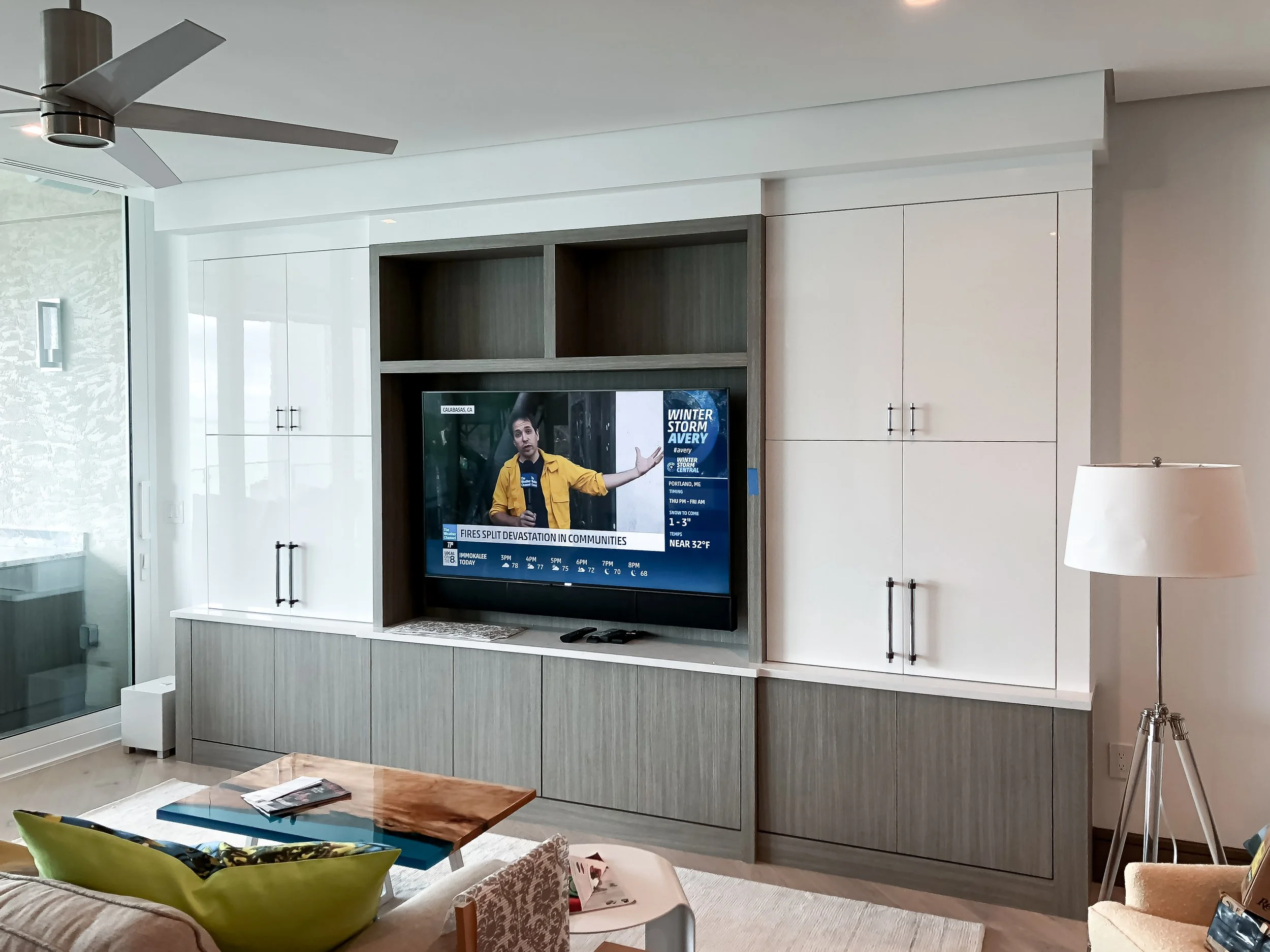 Modern kitchen with white cabinets, a large island with a wooden base and white countertop, two woven pendant lights hanging above, and a large window showing outdoor greenery.