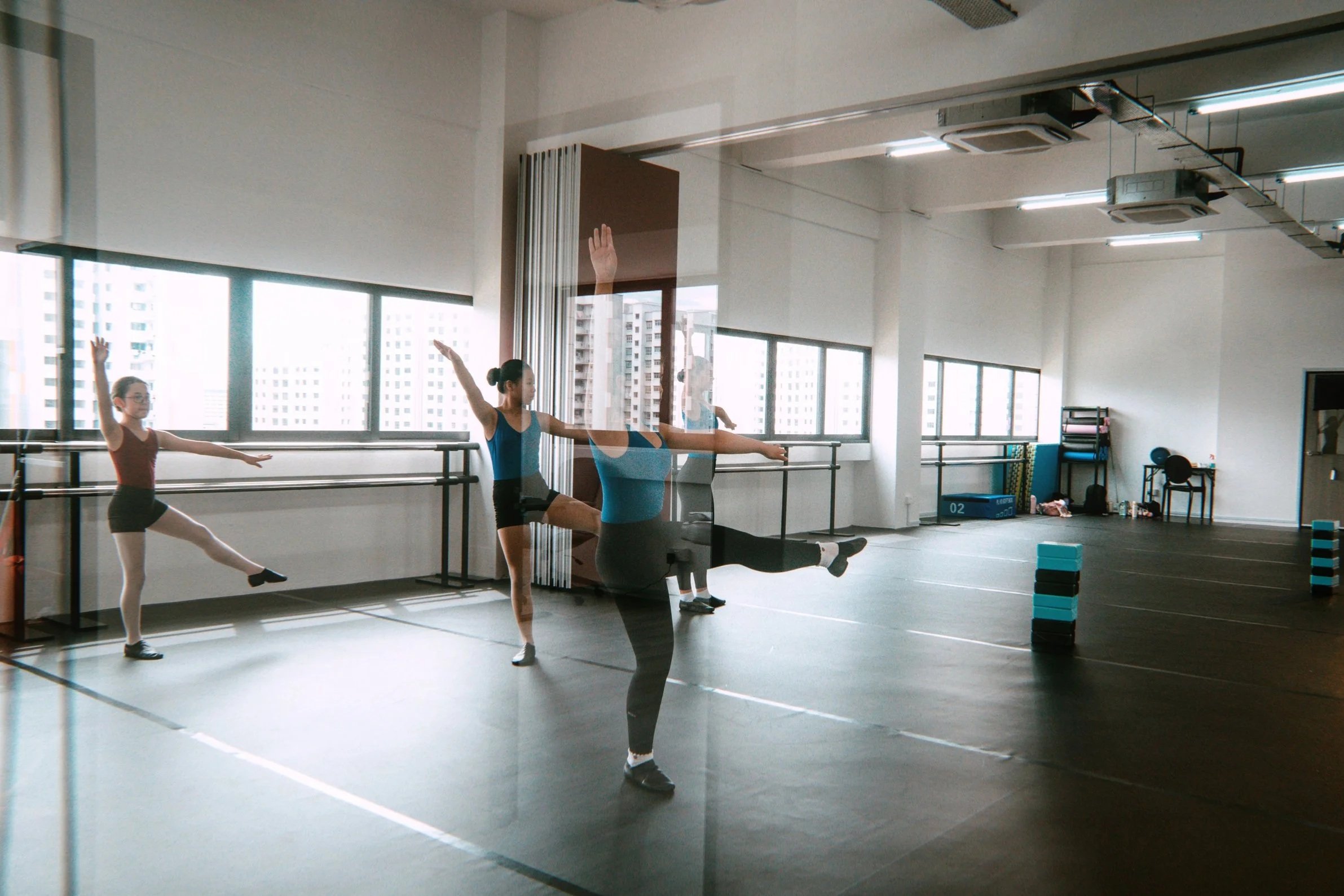 Jazz class in progress with three dancers practicing in a spacious, well-lit studio with large windows.