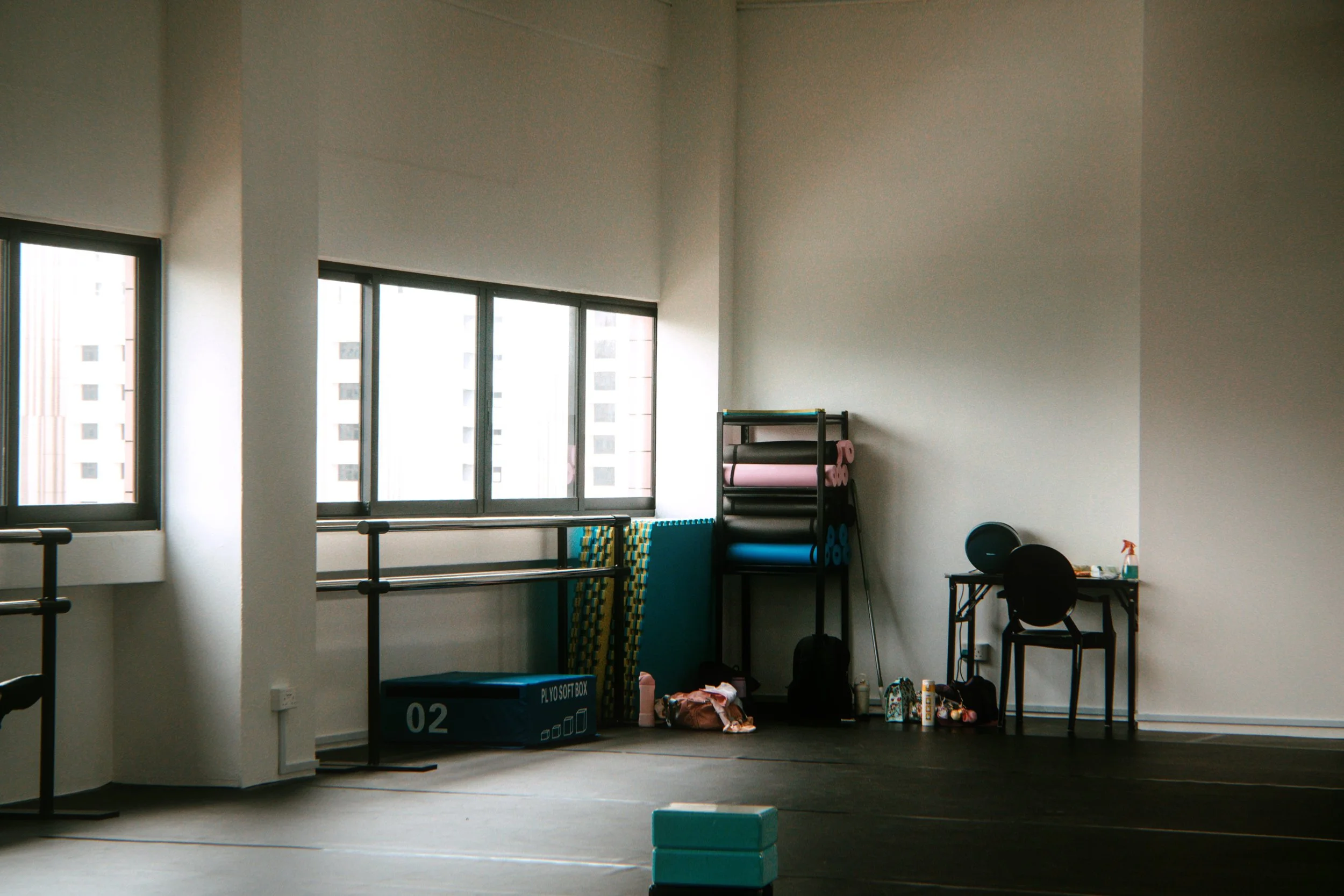 Dance and fitness studio with large windows, workout mats, foam rollers, a balance box, a table with a black chair.