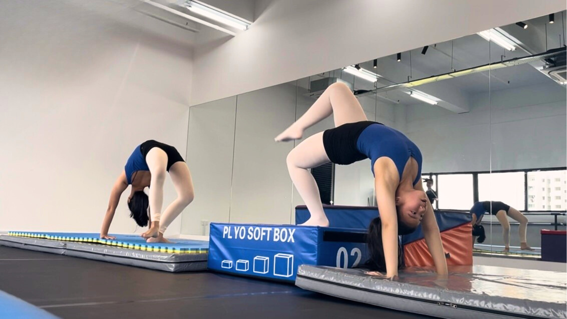 Two young women practicing gymnastics in a dance studio, side by side, one of them upside down and the other in a forward bend, on a padded floor with a soft box support.