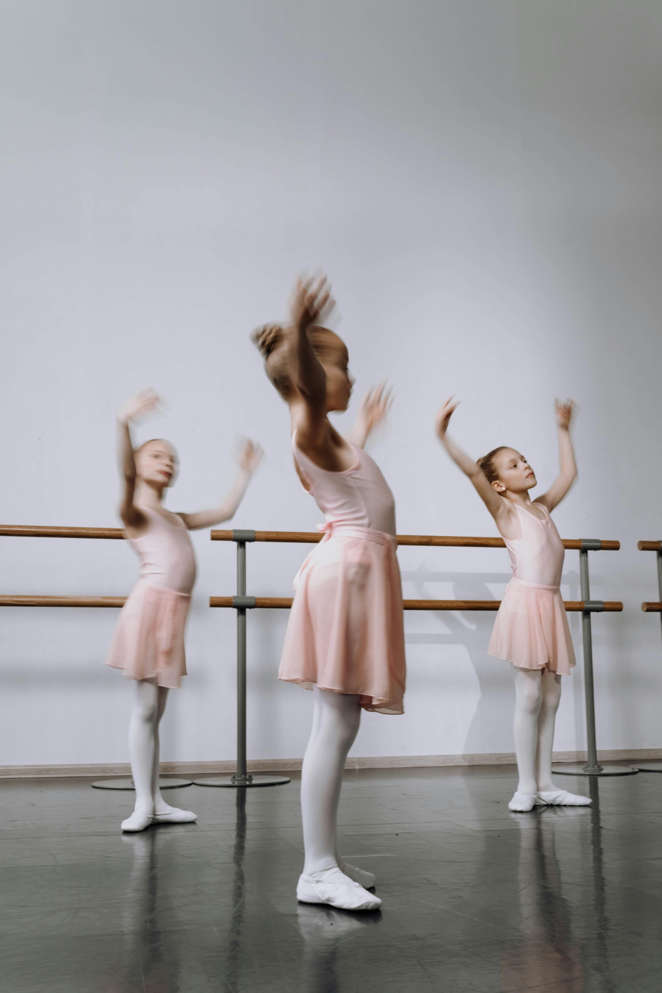 Three young girls in pink ballet dresses and white tights practicing ballet at a dance studio.