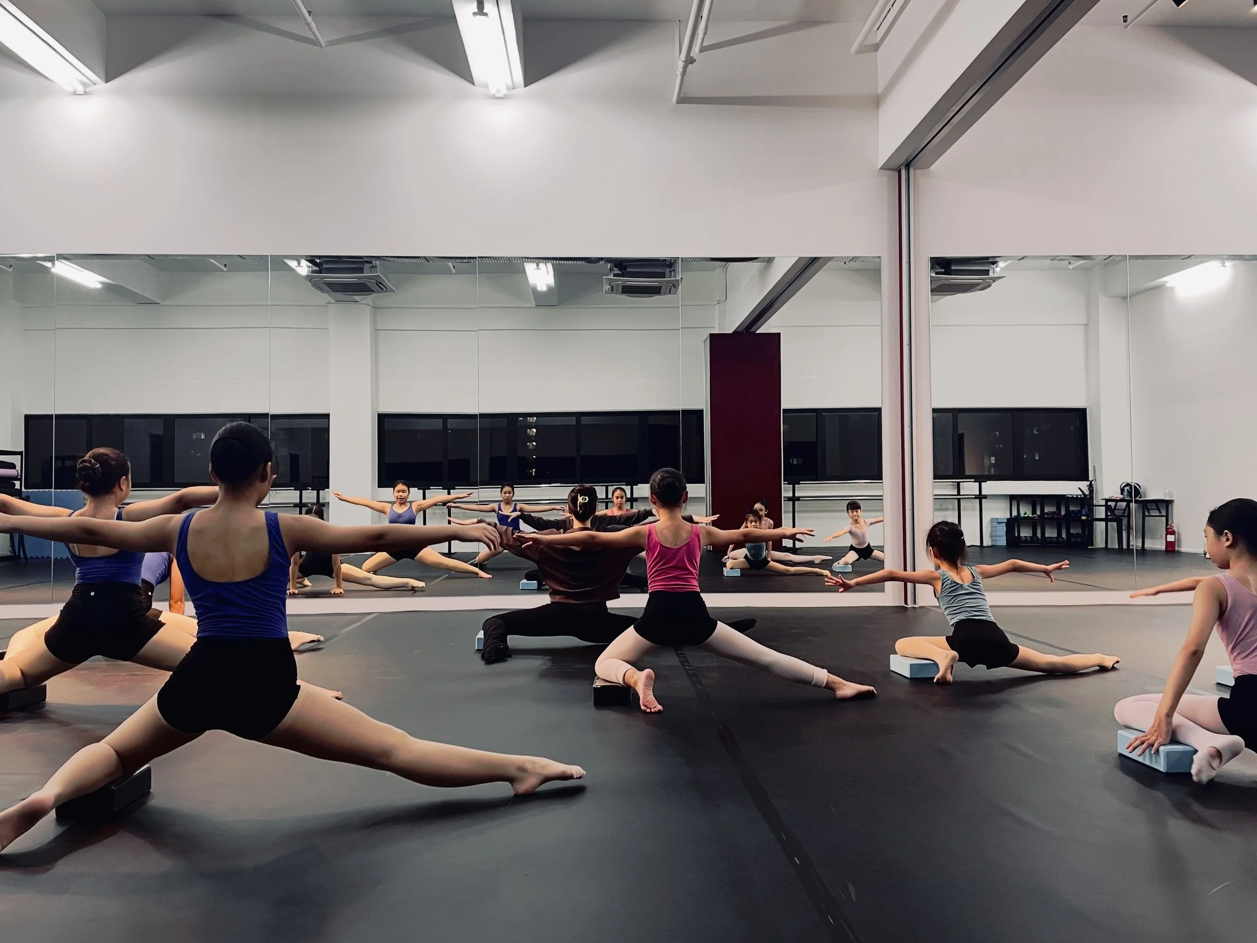 Ballet class with young dancers doing split stretches in a dance studio with mirrors.